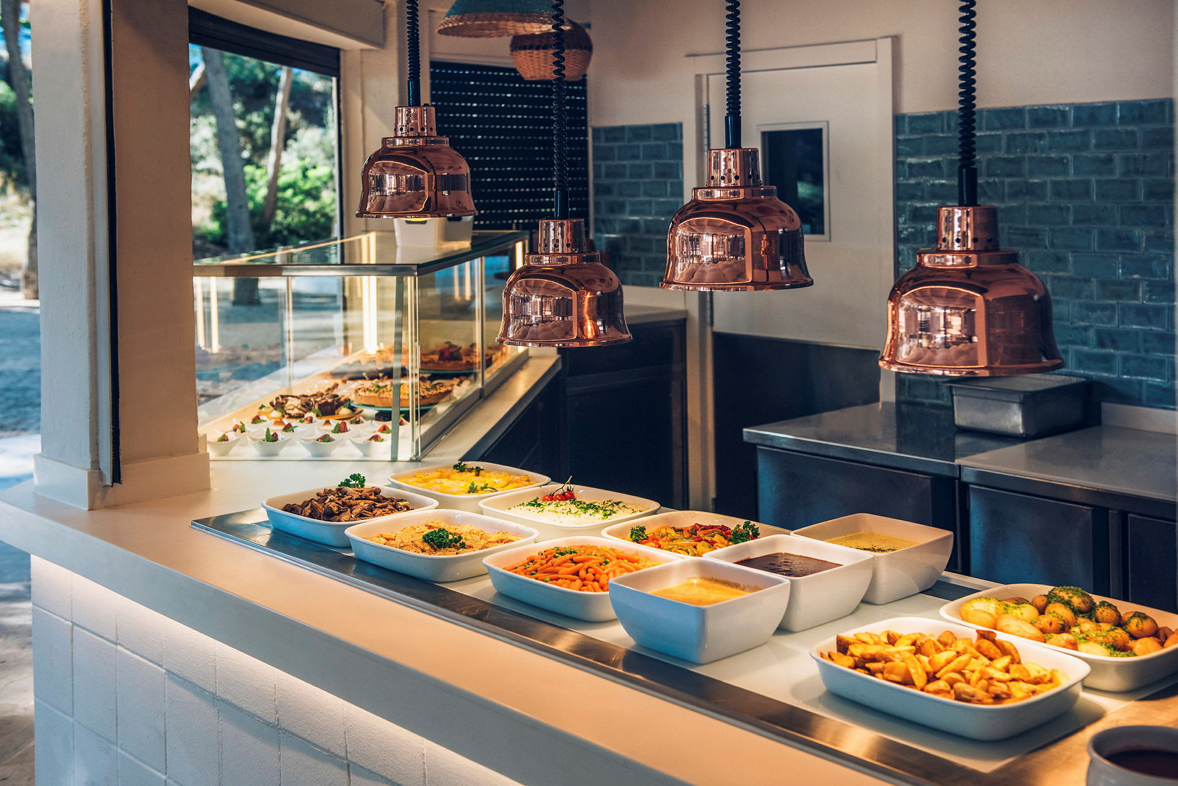 Buffet, jossa useita ruokia tarjoiltuna valkoisissa astioissa tarjoilutiskillä. Yläpuolella on kuparilamppuja. (Tekoälyn luoma teksti)