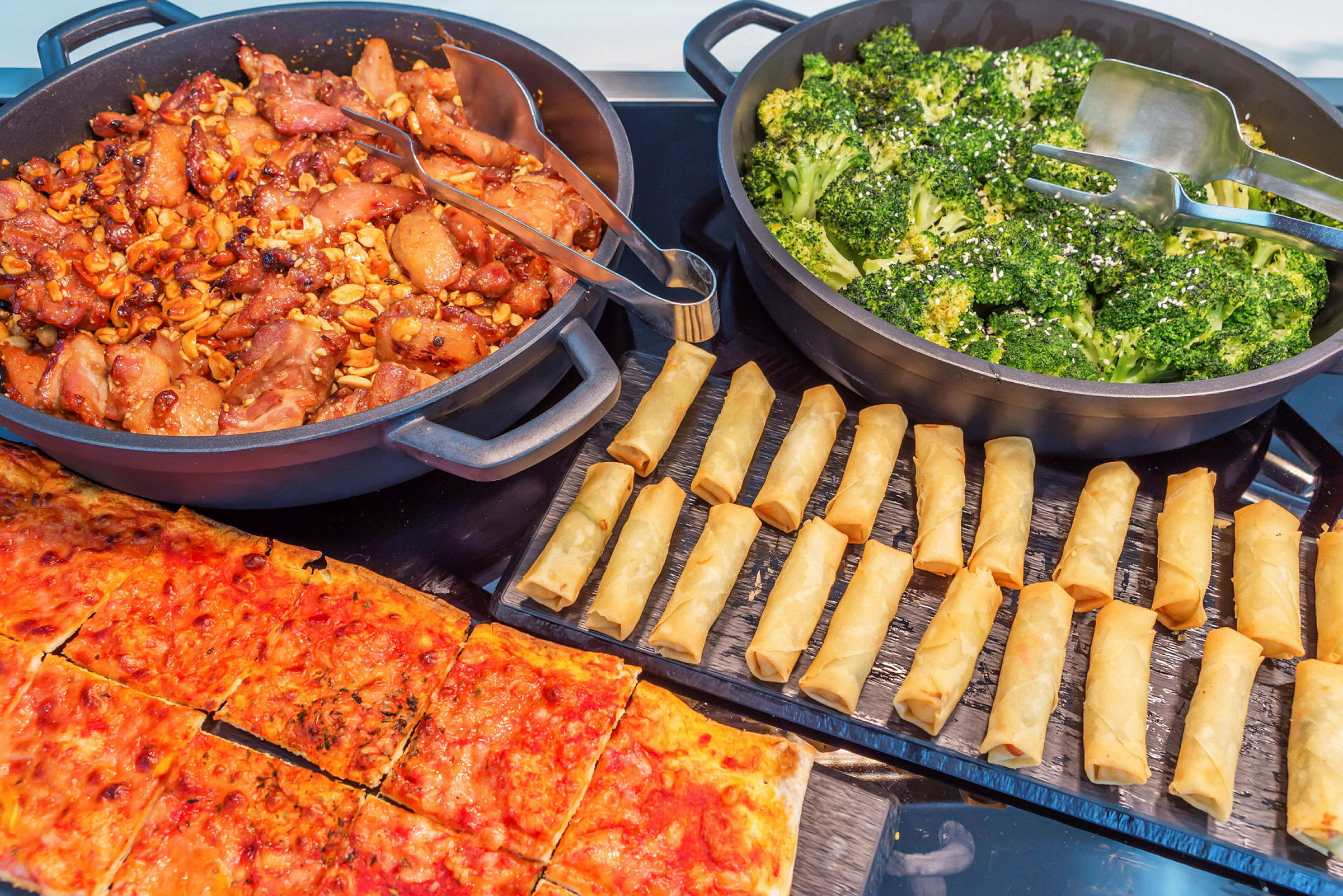 Buffet, jossa erilaisia ruokalajeja, kuten kanaa, parsakaalia, kevätrullia ja pizzaa. (Tekoälyn luoma teksti)