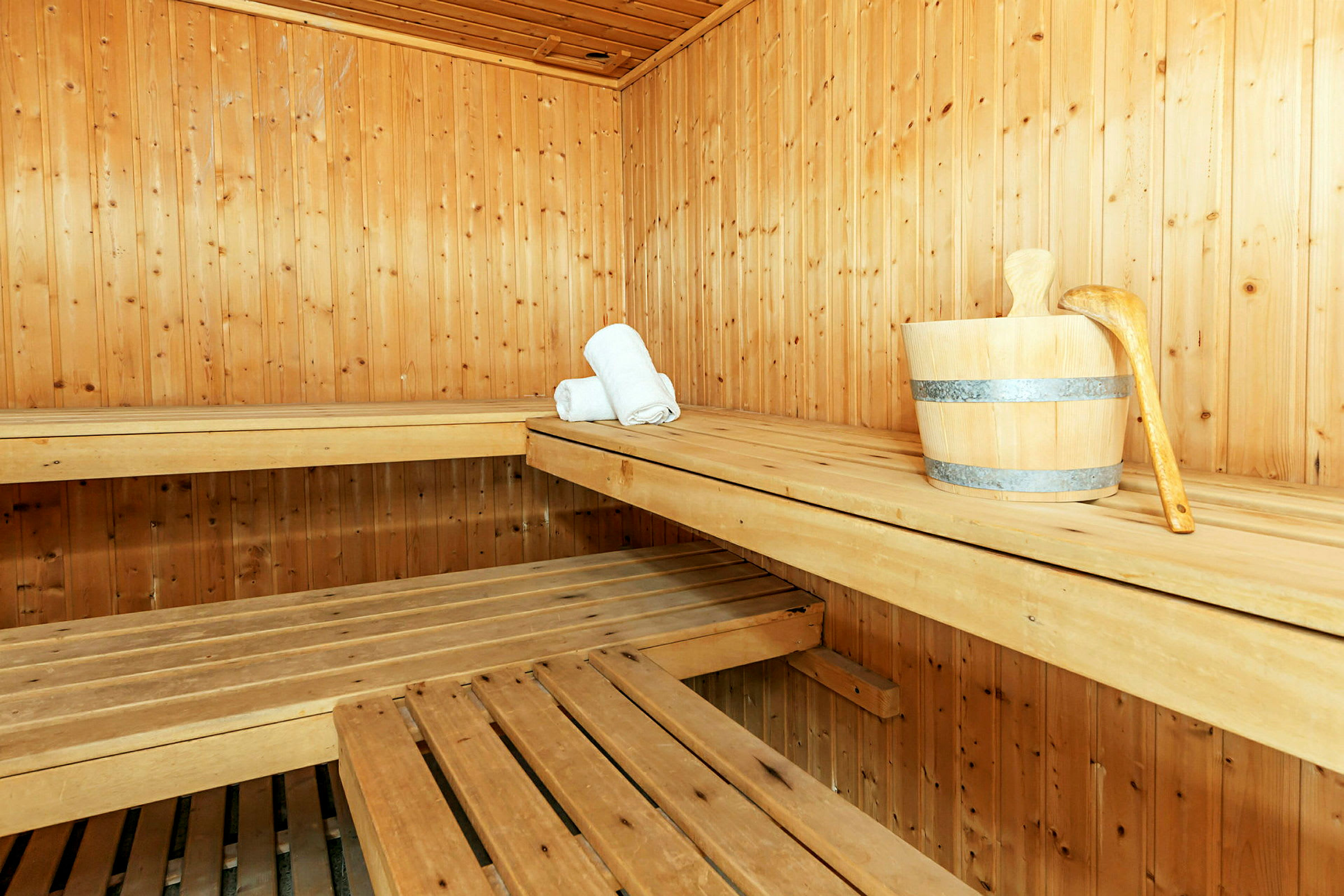 Sauna, jossa puupaneelit, puiset lauteet, puinen sanko kauhalla ja kaksi rullattua valkoista pyyhettä. (Tekoälyn luoma teksti)