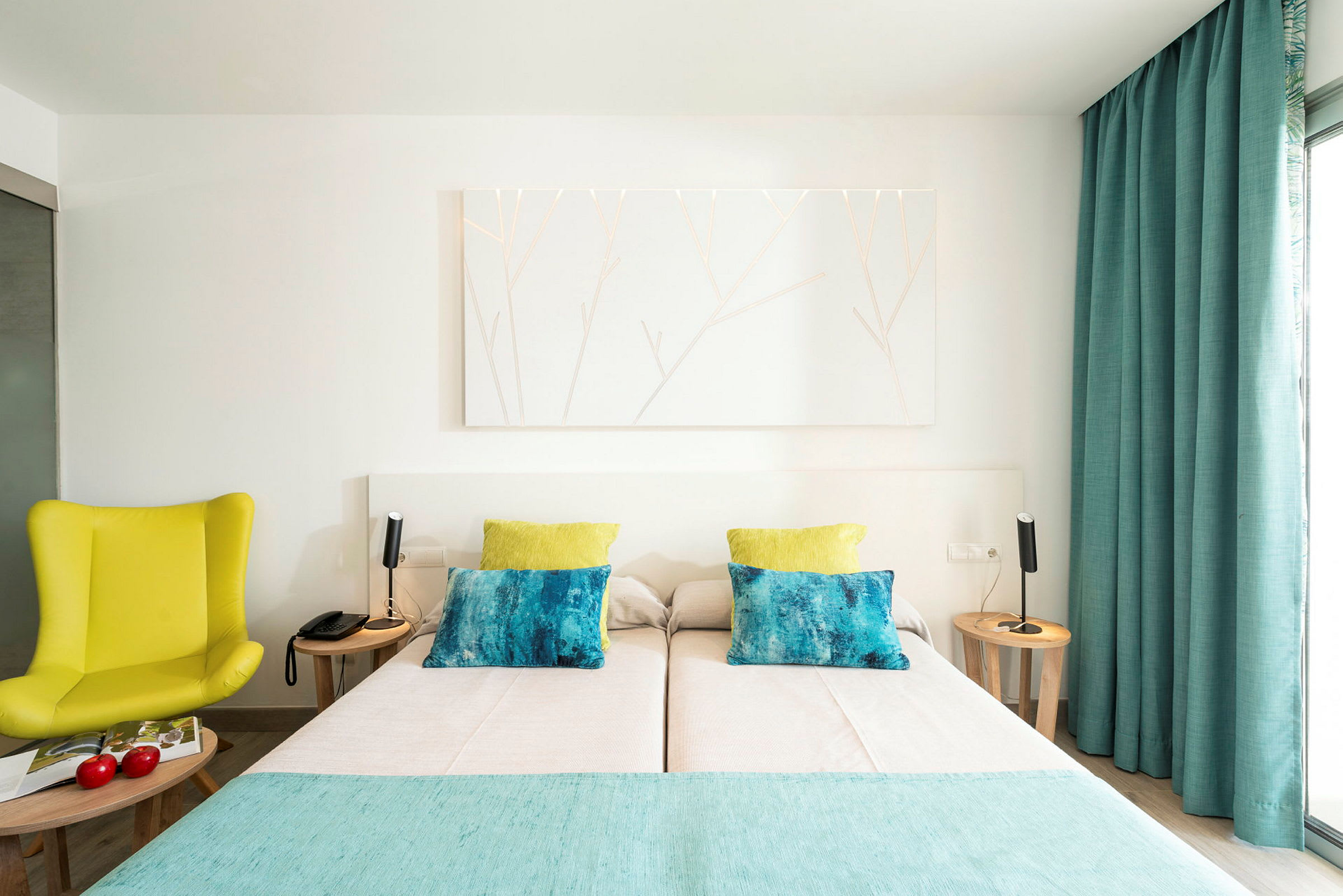 Hotellihuone, jossa on parivuode, kaksi yöpöytää lampuilla, keltainen tuoli ja siniset verhot. (Tekoälyn luoma teksti)