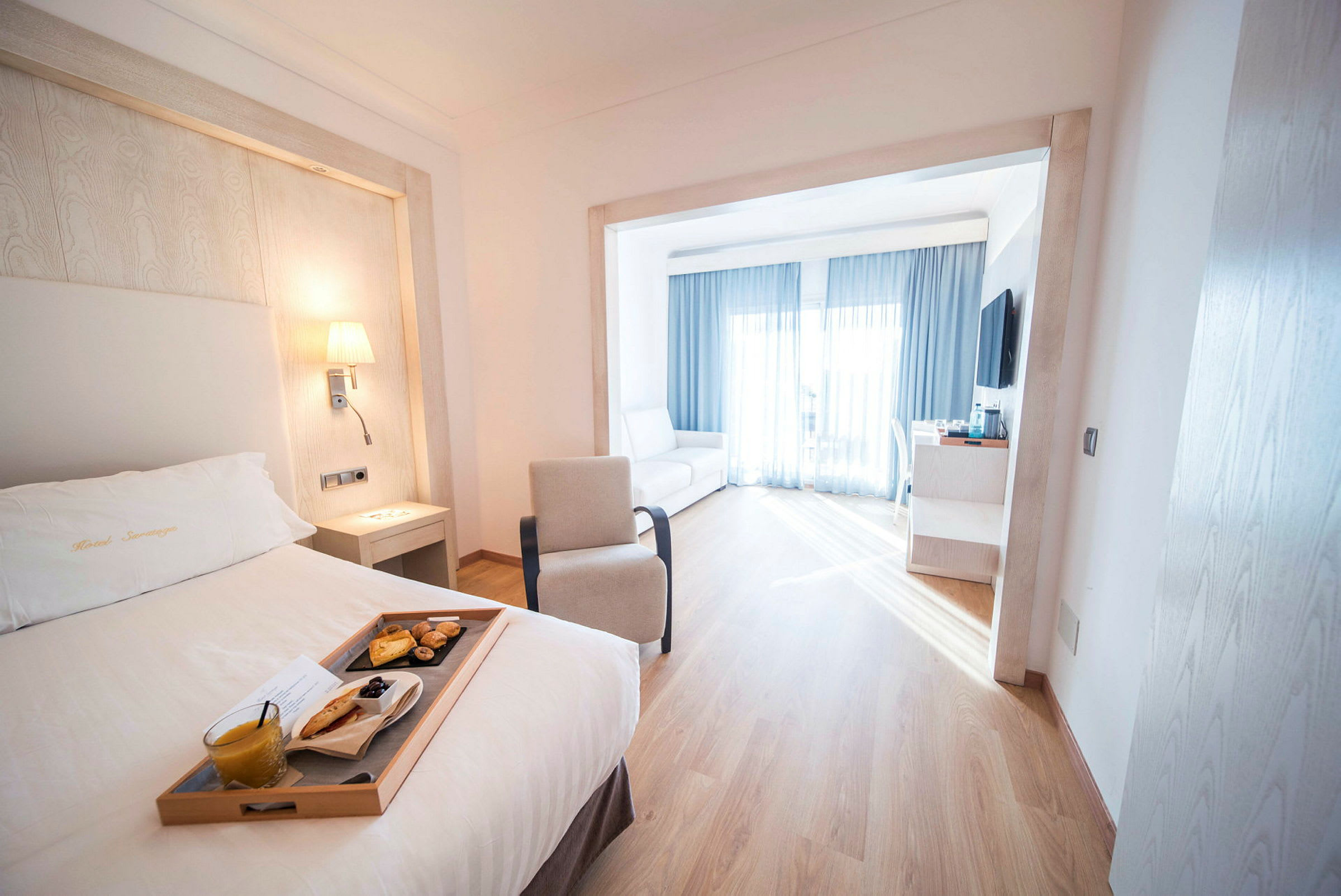 Hotellihuone, jossa on sänky ja tarjotin, joka sisältää aamiaista. Huoneessa on iso ikkuna, jossa on vaaleat verhot. (Tekoälyn luoma teksti)