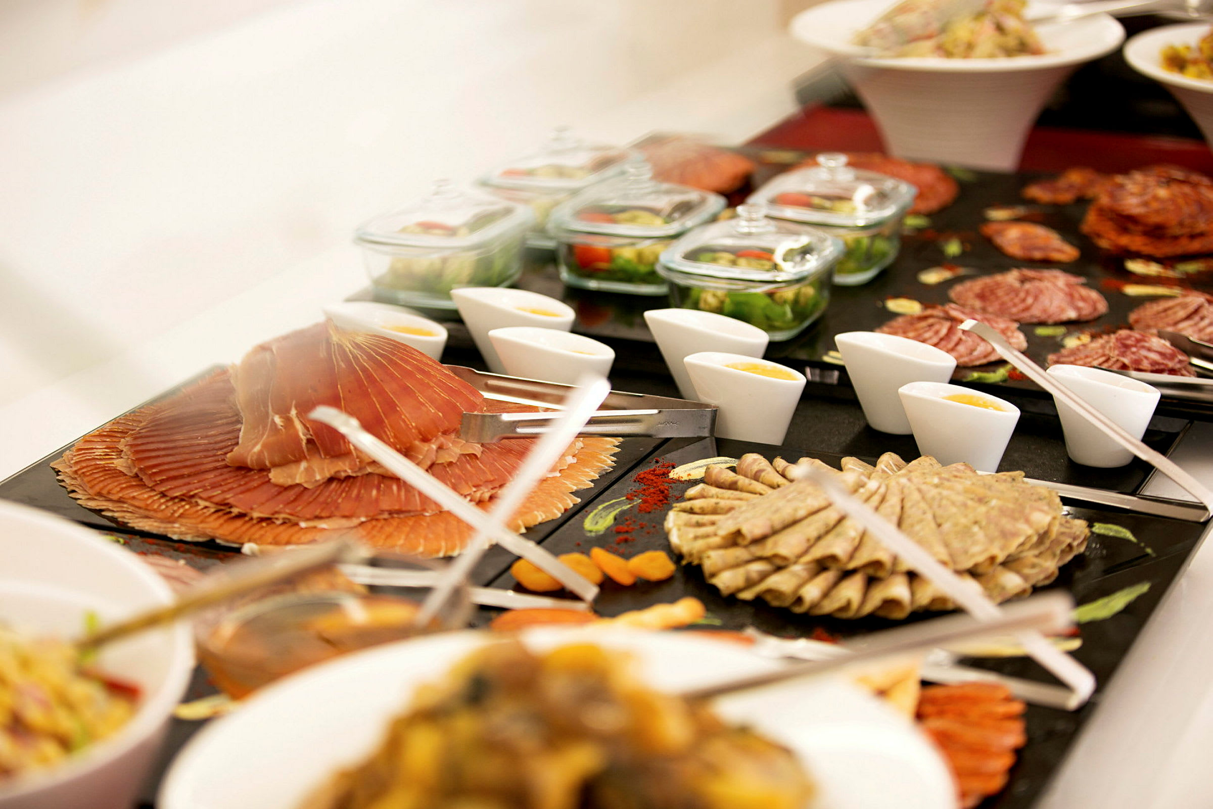 Buffet, jossa on erilaisia leikkeleitä, salaatteja, kastikkeita ja lisukkeita esitettyinä kulhoissa ja lautasilla. (Tekoälyn luoma teksti)