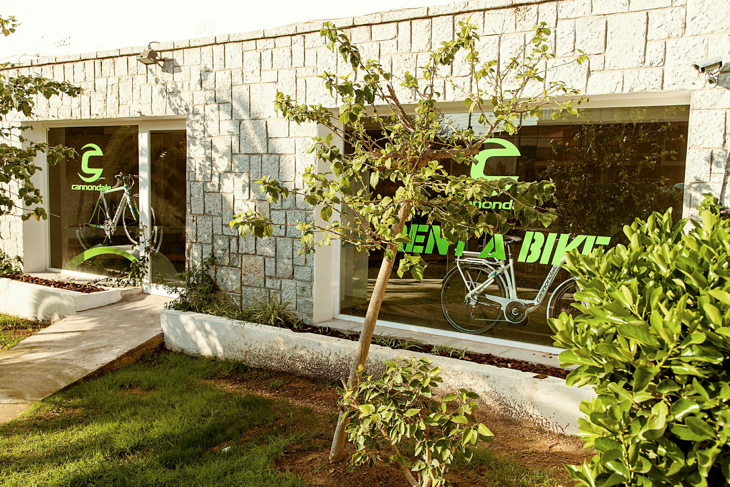 Rakennus, jossa on kiviseinät ja suuret lasi-ikkunat. Ikkunoissa on teksti "Cannondale Rent a Bike" ja kuva pyörästä. Rakennuksen edessä kasvaa puita ja pensaita. (Tekoälyn luoma teksti)