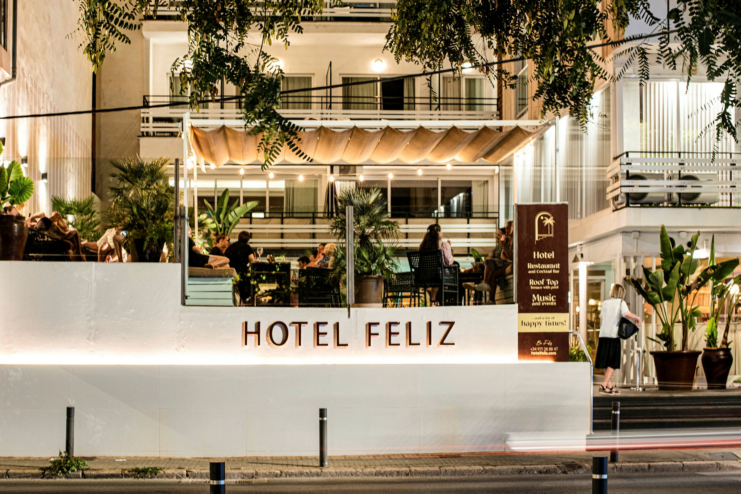 Hotel Feliz, jossa on valaistu julkisivu ja terassi, jossa ihmiset istuvat. (Tekoälyn luoma teksti)