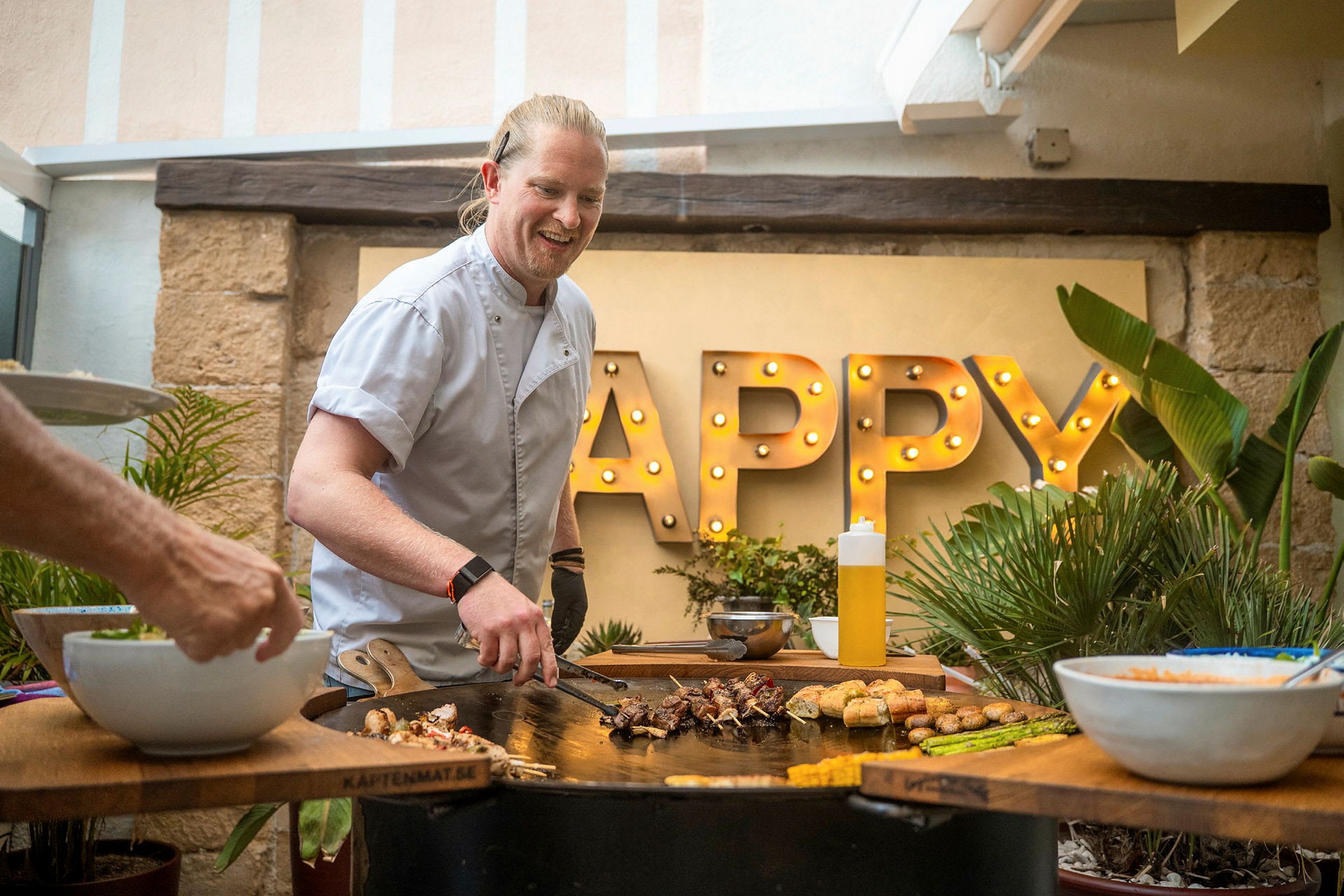 Henkilö valmistaa ruokaa grillillä, taustalla valaistu sana 'Happy'. (Tekoälyn luoma teksti)