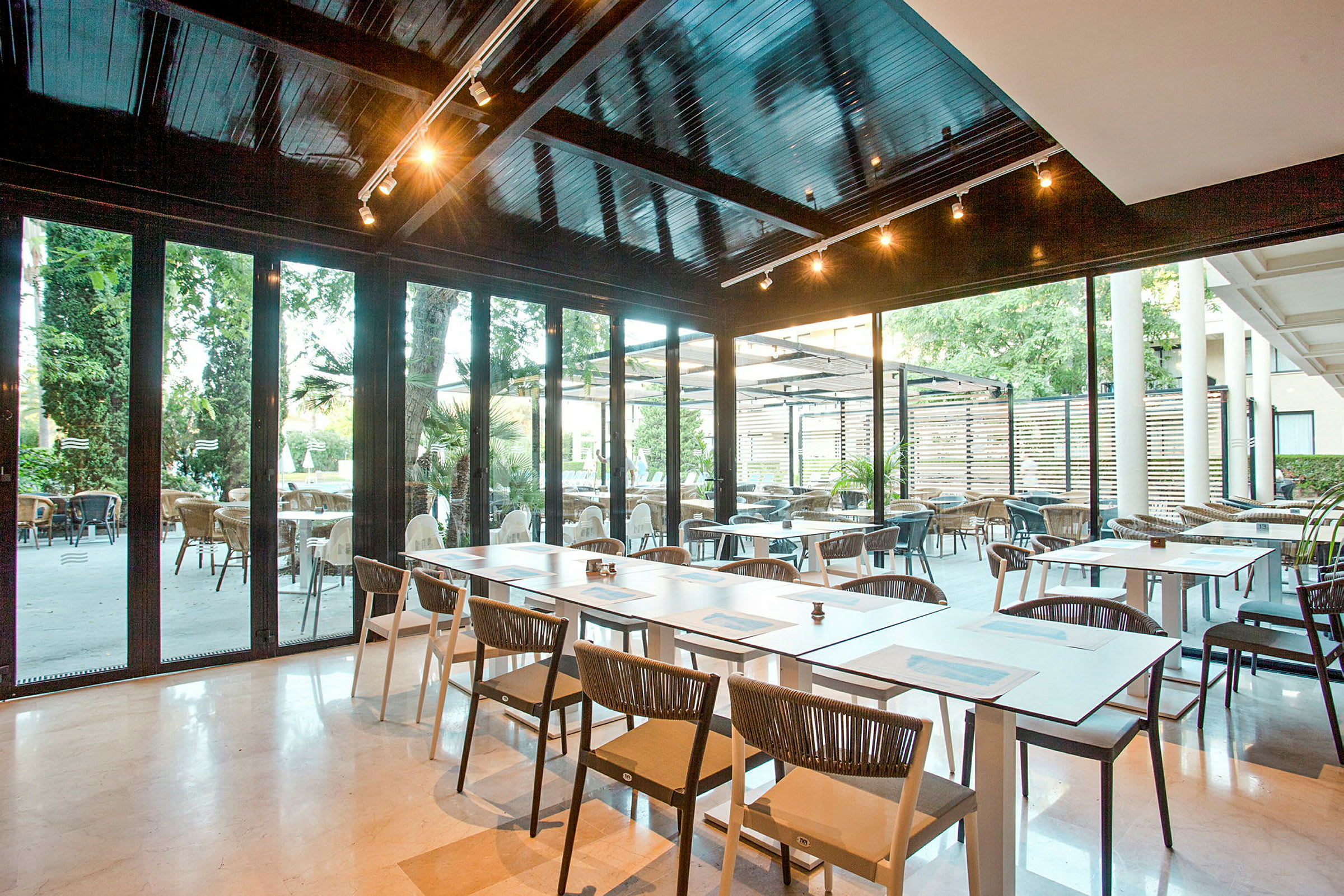 Moderni ravintola, jossa on istumapaikkoja sisä- ja ulkotiloissa. Pöydät ja tuolit on sijoitettu vaalealle lattialle, ja lasiseinät tarjoavat näkymän ulkoalueelle. (Tekoälyn luoma teksti)