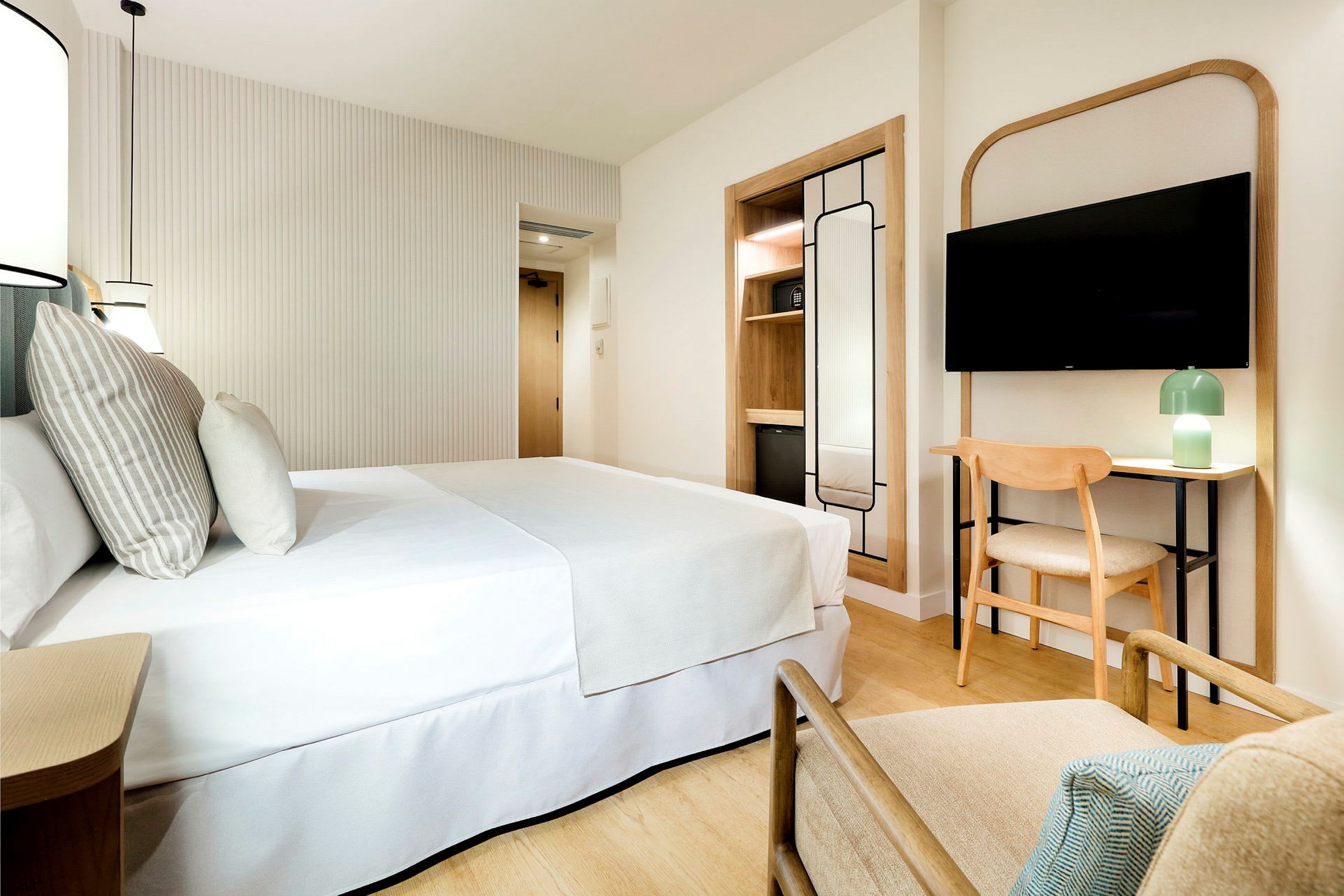 Moderni ja valoisa hotellihuone, jossa on parivuode, seinään kiinnitetty TV, nojatuoli, työpöytä tuoleineen ja minimalistinen sisustus. (Tekoälyn luoma teksti)