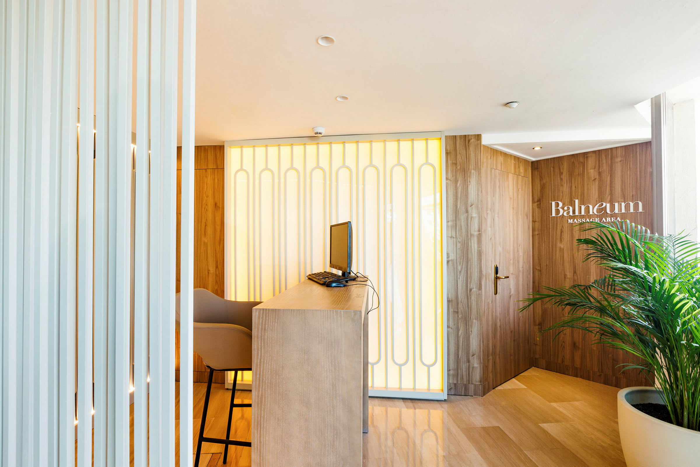 Vastaanotto, jossa on pöytä, tietokone ja tuoli, taustalla seinäpaneelit ja ovi, jossa lukee 'Balneum Massage Area'. (Tekoälyn luoma teksti)