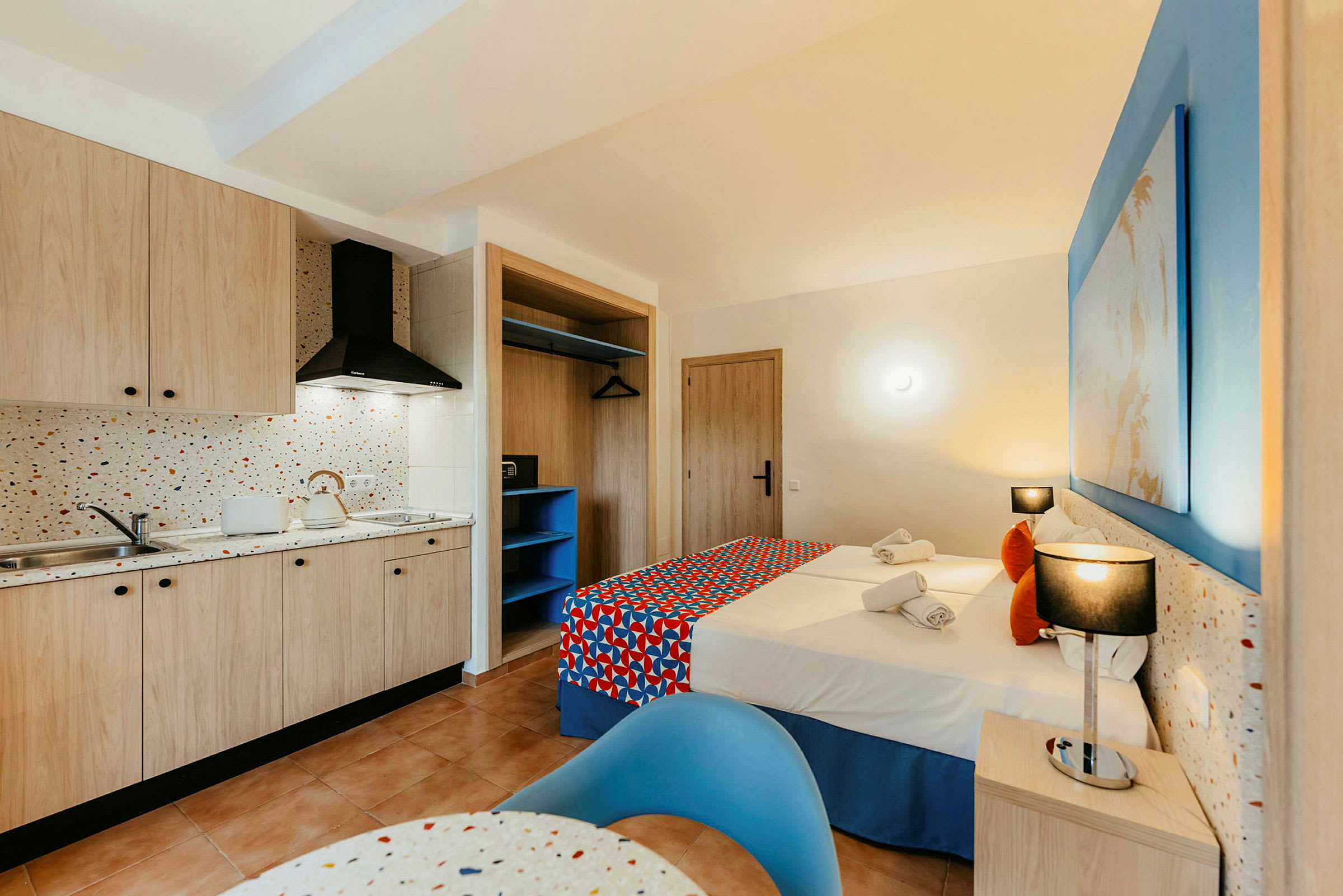 Hotellihuone, jossa on keittiö ja makuutila. Keittiössä puukaapit, tiskiallas ja liesi. Makuutilassa kaksi sänkyä värikkäillä päiväpeitteillä, yöpöytä ja valaistus. (Tekoälyn luoma teksti)