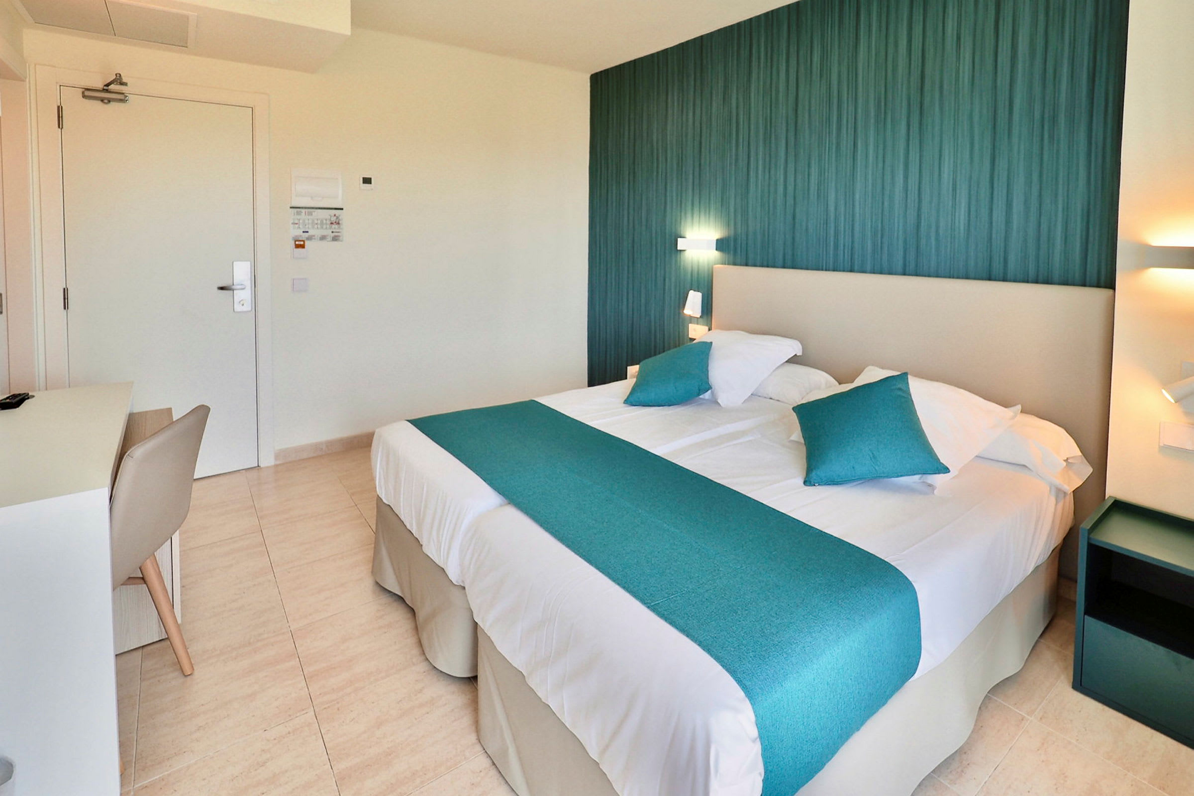 Hotellihuone, jossa on parivuode ja siniset koristeelliset tyynyt sekä päiväpeite. Vuoteen vieressä näkyy lamppu ja yöpöytä. (Tekoälyn luoma teksti)