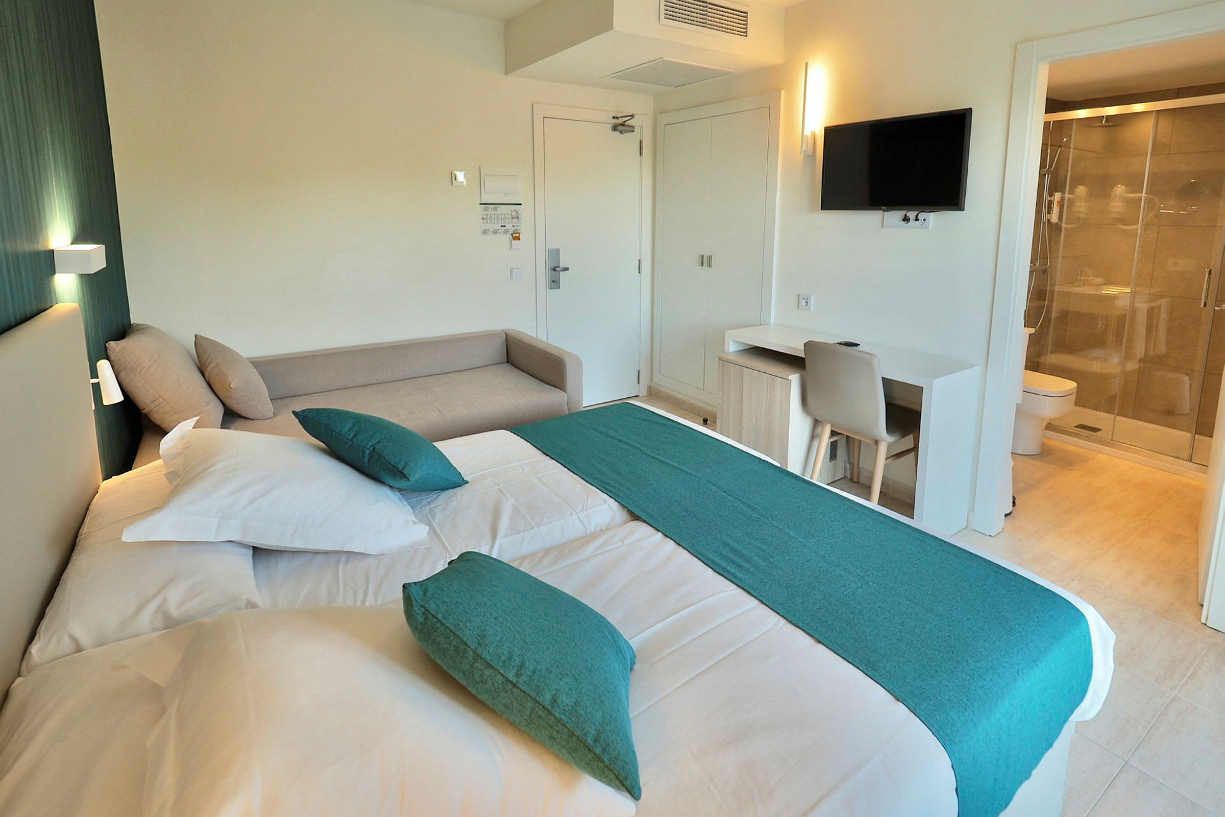 Hotellihuone, jossa on sänky, valkoiset lakanat, koristeelliset vihreät tyynyt ja yhteensopiva vihreä peite. Huoneessa on vaalea sohva, kirjoituspöytä tuoleineen ja seinälle kiinnitetty televisio. Kylpyhuone, jossa on suihku, näkyy oikealla lasioven takaa. (Tekoälyn luoma teksti)
