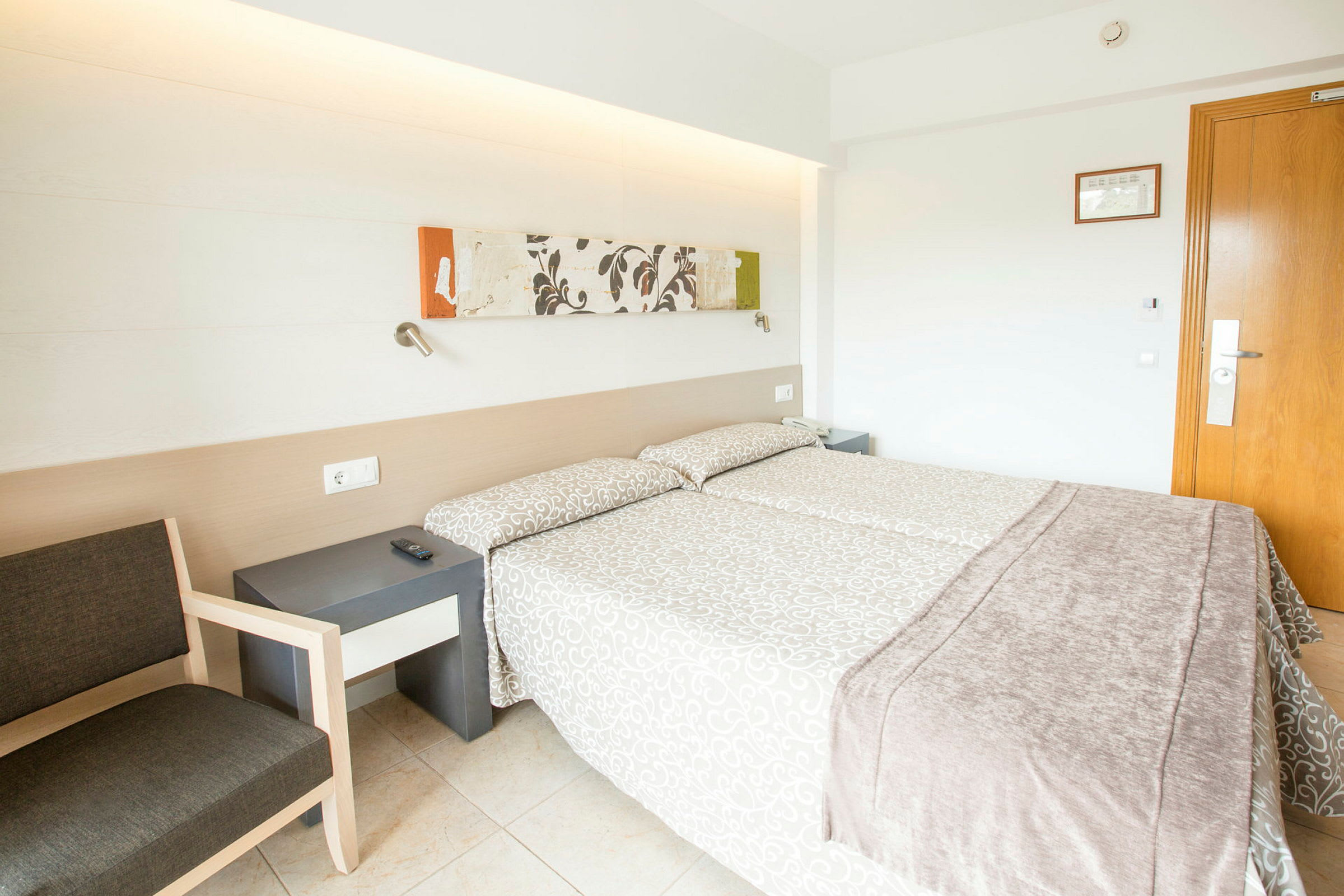 Hotellihuone, jossa on kaksi sänkyä, yöpöytä ja tuoli. Seinän koristelu ja ovi näkyvät taustalla. (Tekoälyn luoma teksti)