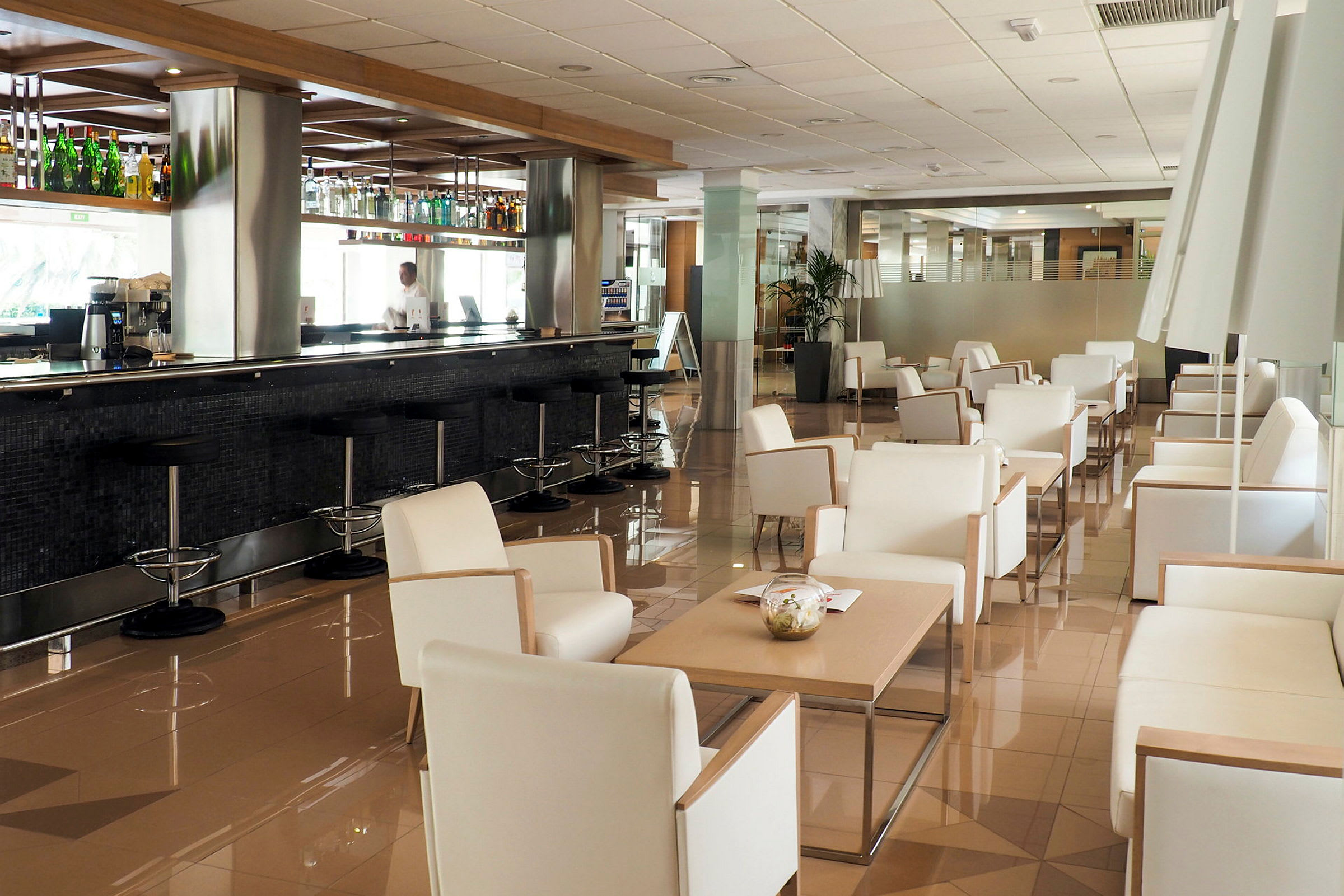 Moderni lounge-alue, jossa valkoiset nojatuolit ja pöydät sekä pitkä baaritiski tuoleineen. (Tekoälyn luoma teksti)