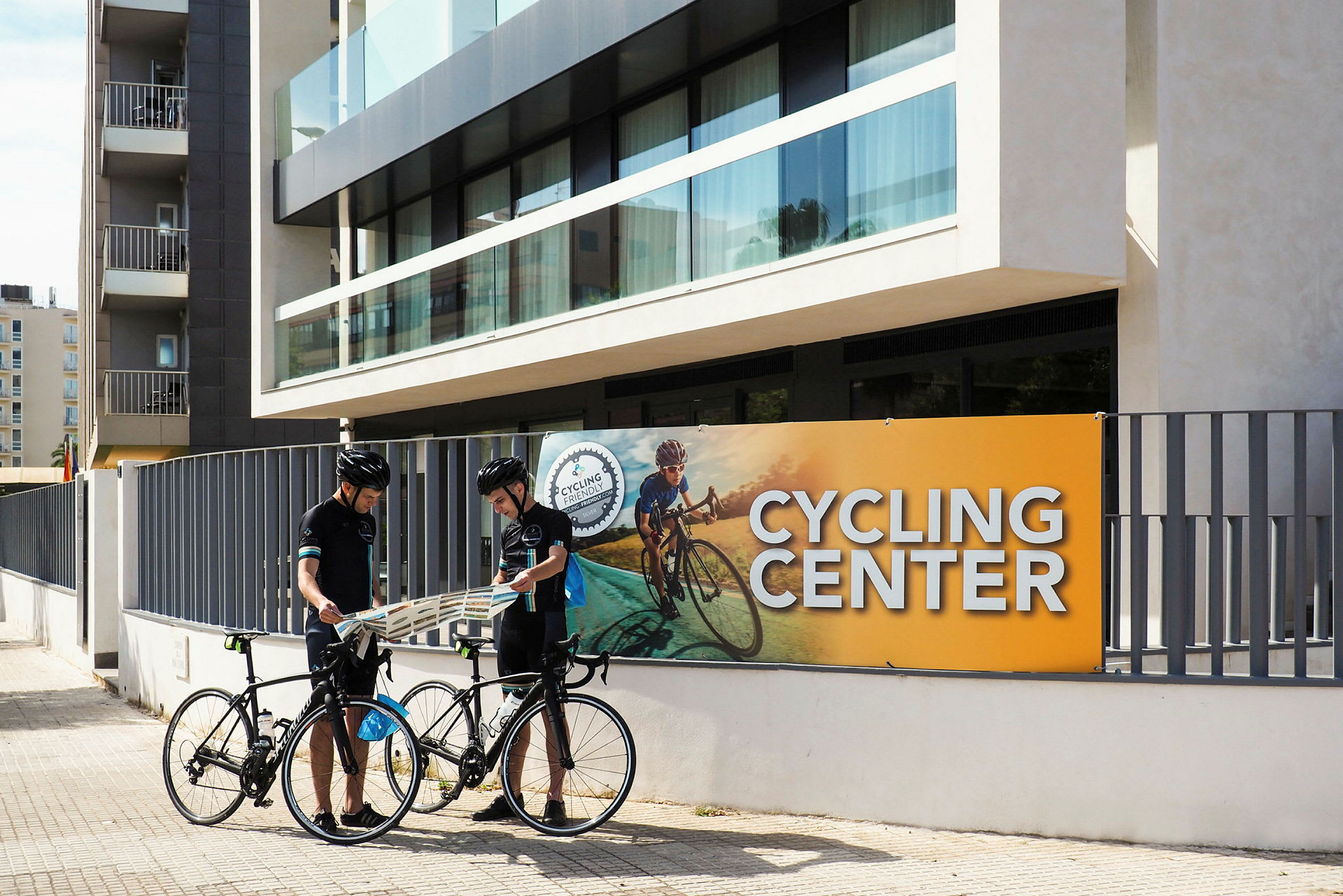 Kaksi henkilöä seisoo polkupyöriensä kanssa rakennuksen edessä, jossa lukee Cycling Center. (Tekoälyn luoma teksti)