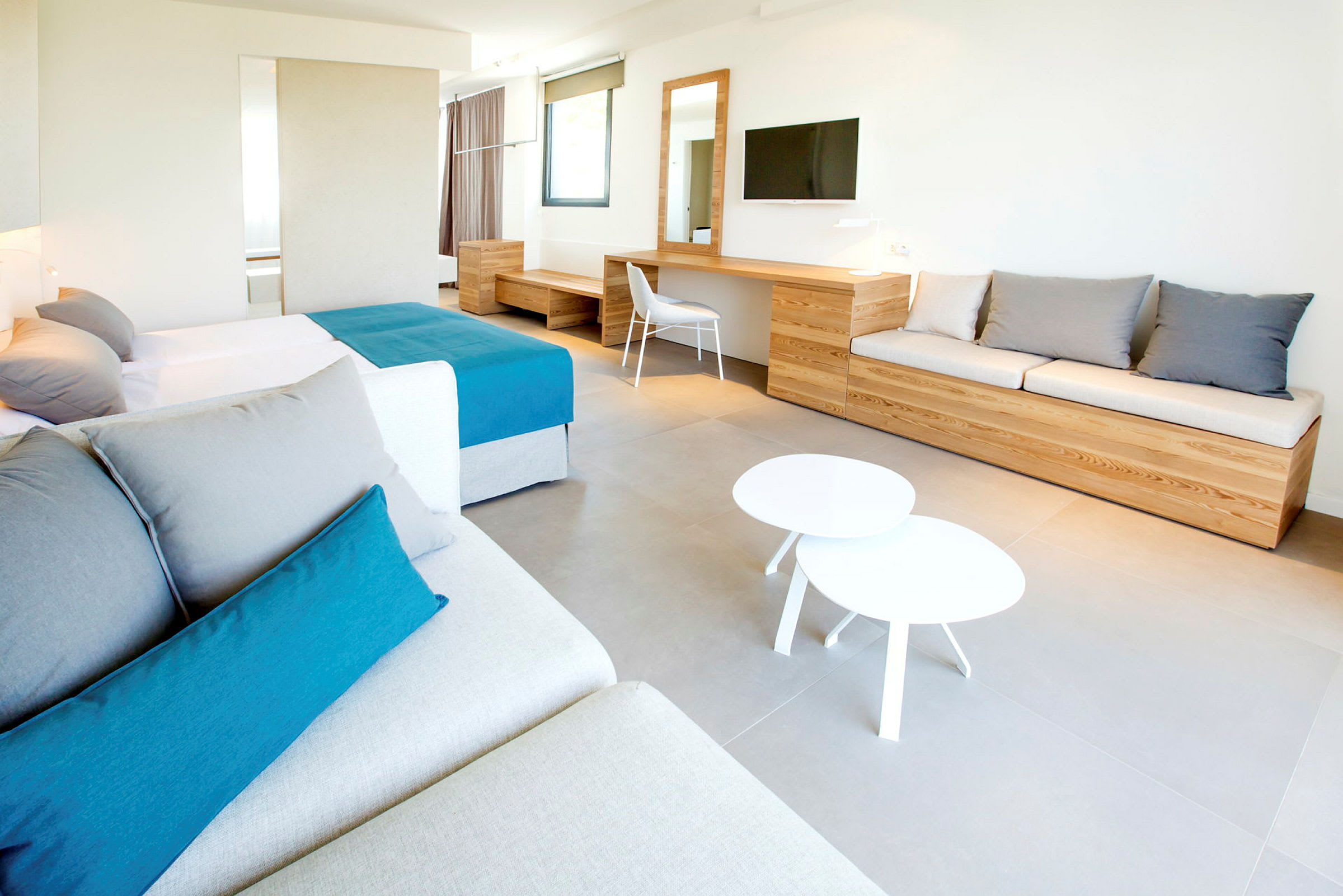 Moderni hotellihuone, jossa on iso sänky, sohva, työpöytä ja seinään kiinnitetty televisio. Huonekalut ovat puunvärisiä, ja huoneen tyyli on minimalistinen. (Tekoälyn luoma teksti)