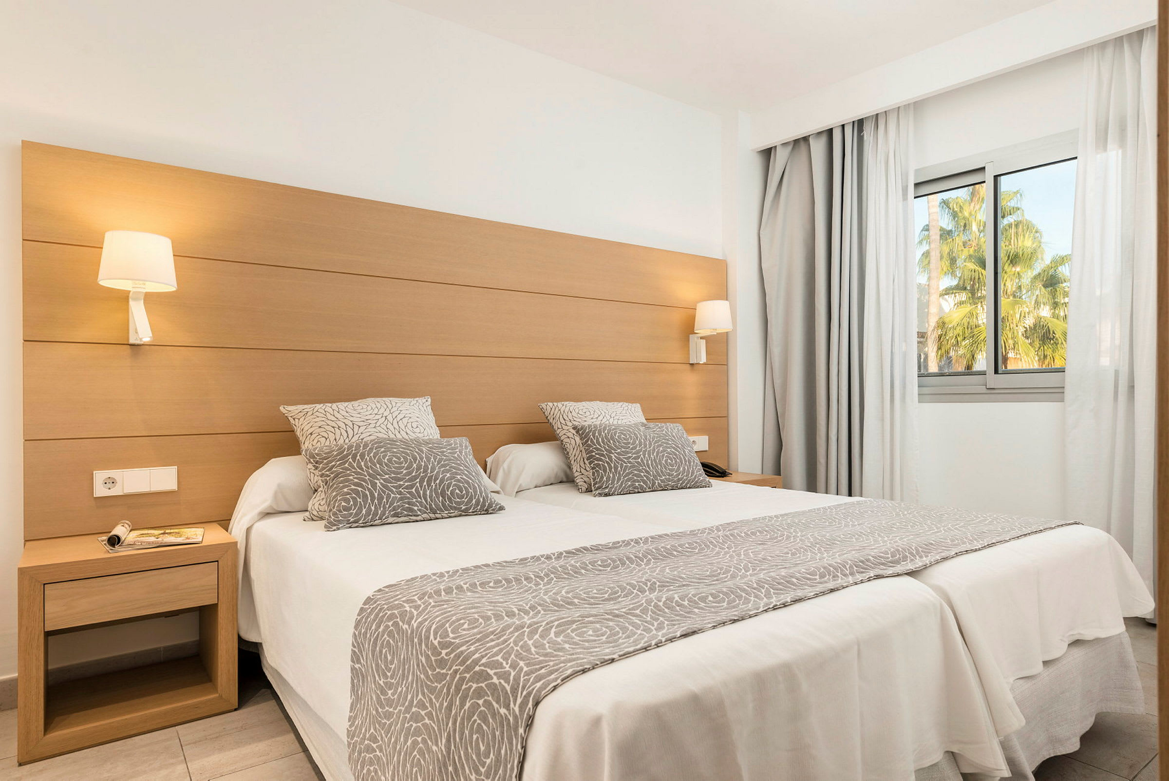 Hotellihuone, jossa on kaksi yhdistettyä yhden hengen vuodetta, koristeltu harmaa-valkoisilla kuvioituilla tekstiileillä, ja iso ikkuna näkymällä vehreyteen. (Tekoälyn luoma teksti)