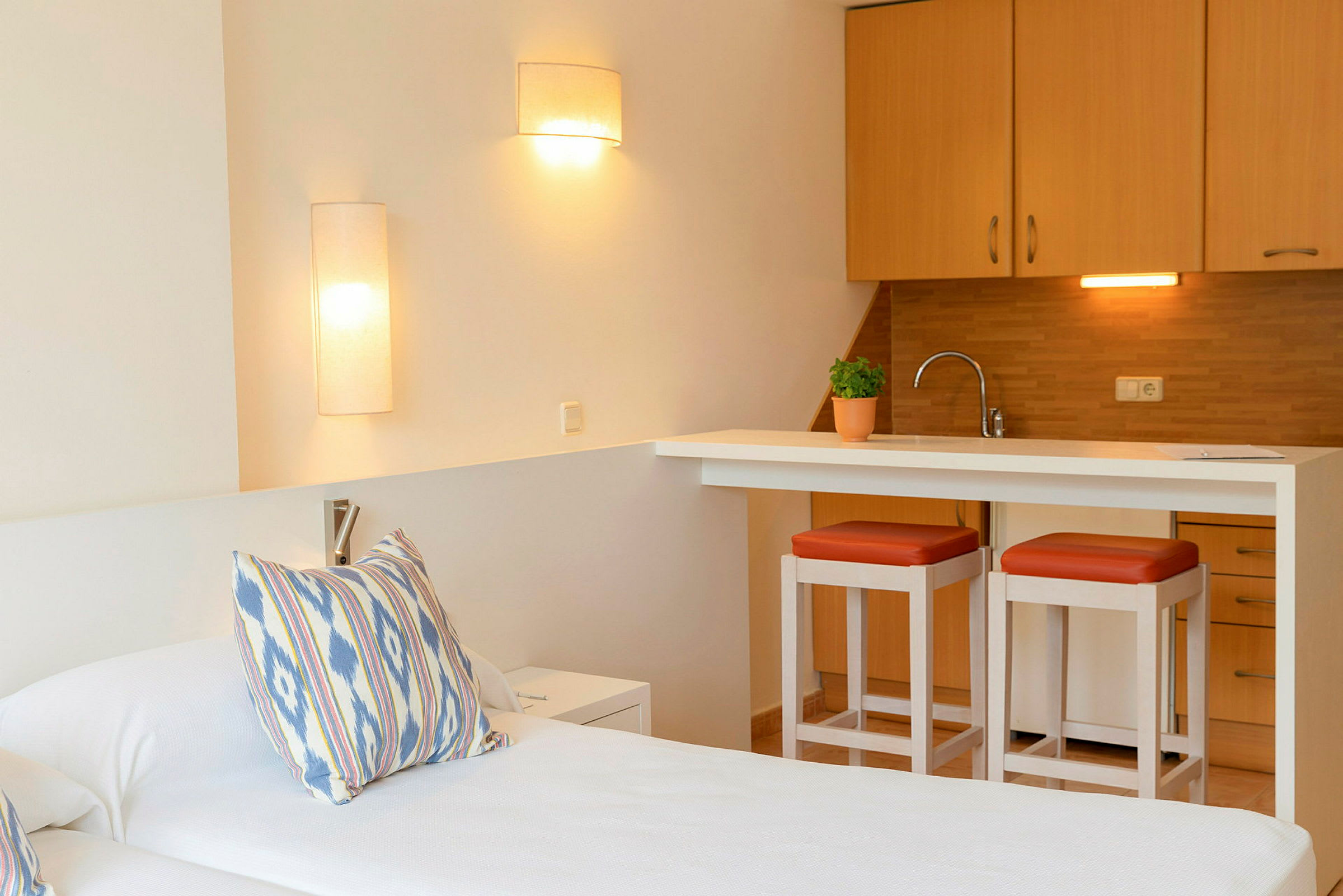 Hotellihuone, jossa on yksinkertainen sisustus, yksi sänky, tyynyjä, pieni keittiötila baarituoleilla ja kaapeilla. (Tekoälyn luoma teksti)