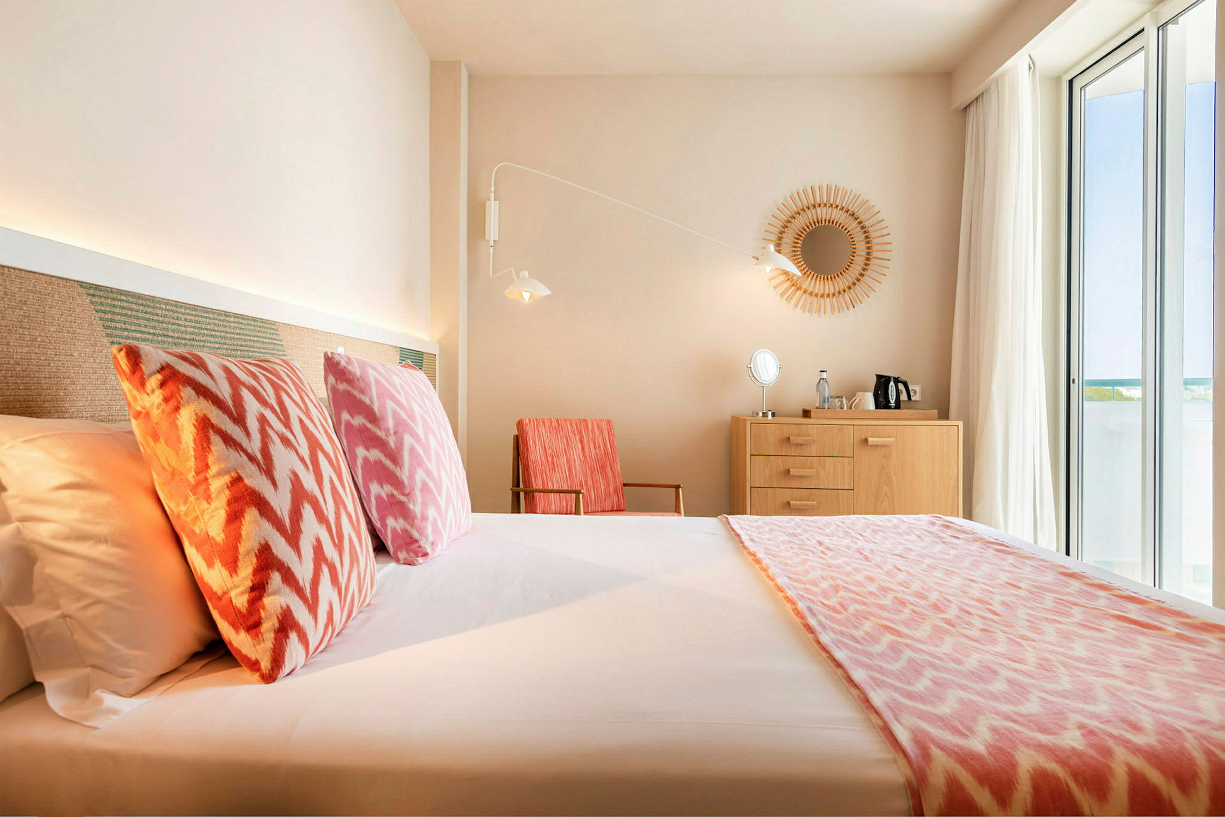 Hotellihuone, jossa on sänky, värikkäät tyynyt, puinen lipasto ja suuret ikkunat. (Tekoälyn luoma teksti)