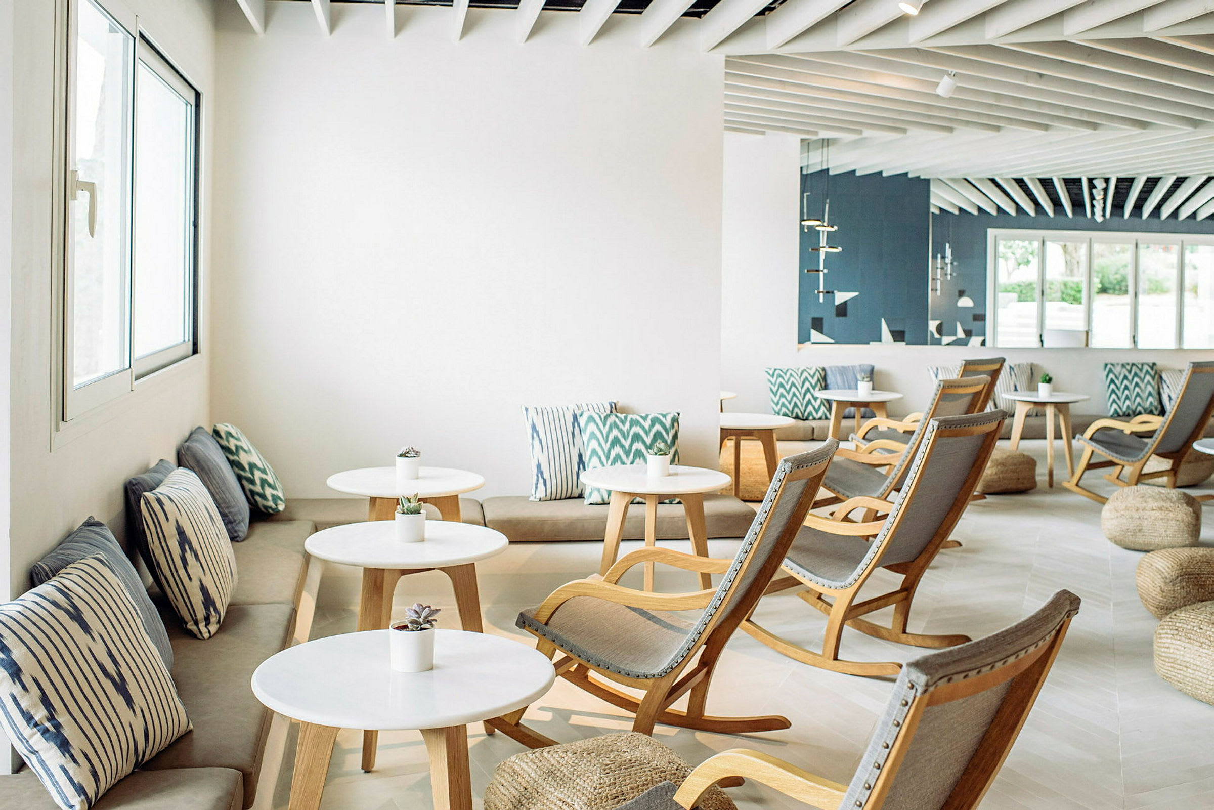 Valoisa lounge-tila, jossa on valkoisia pöytiä, puutuoleja ja sohvia, koristeltu kuvioiduilla tyynyillä. (Tekoälyn luoma teksti)
