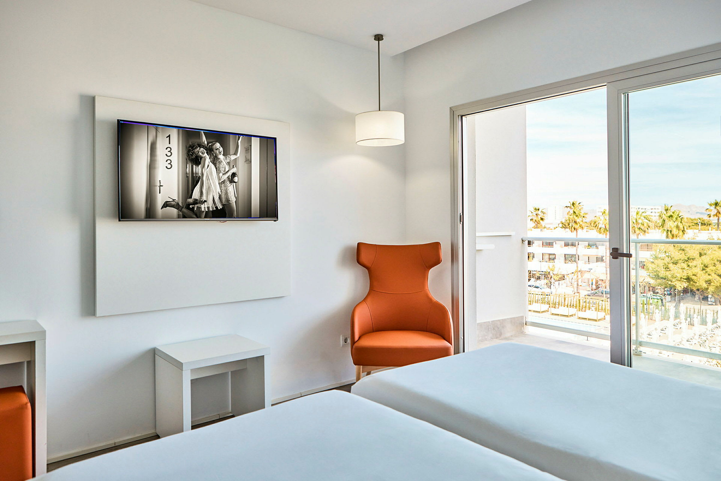Hotellihuone, jossa on kaksi yhden hengen vuodetta, oranssi nojatuoli ja seinälle asennettu televisio. Ikkunan kautta näkyy parveke ja ulkotilaa. (Tekoälyn luoma teksti)
