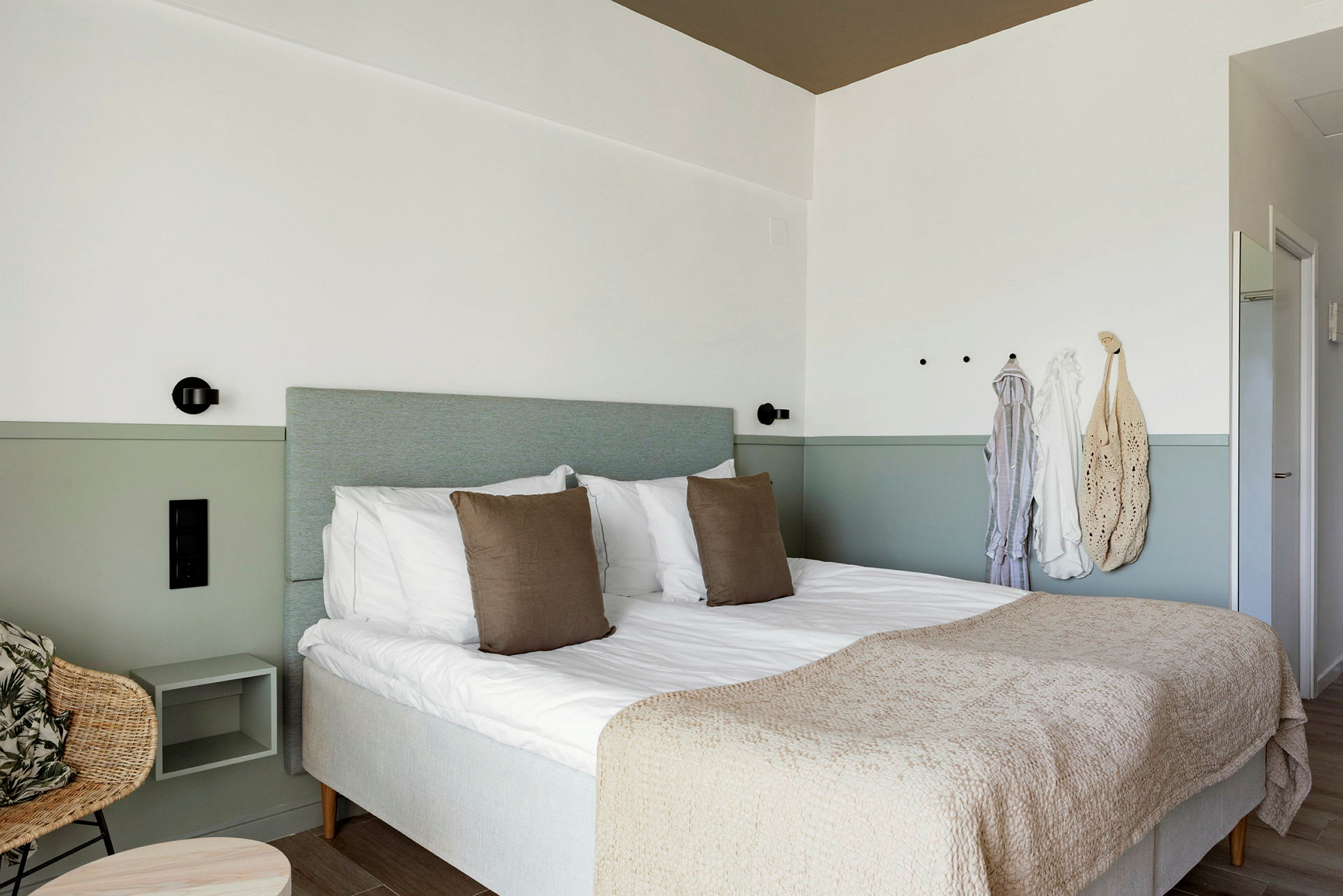 Hotellihuone, jossa on parivuode, kaksi tyynyä ja beige sängynpeite. Seinällä koukkuja ja roikkuva vaatetus. (Tekoälyn luoma teksti)