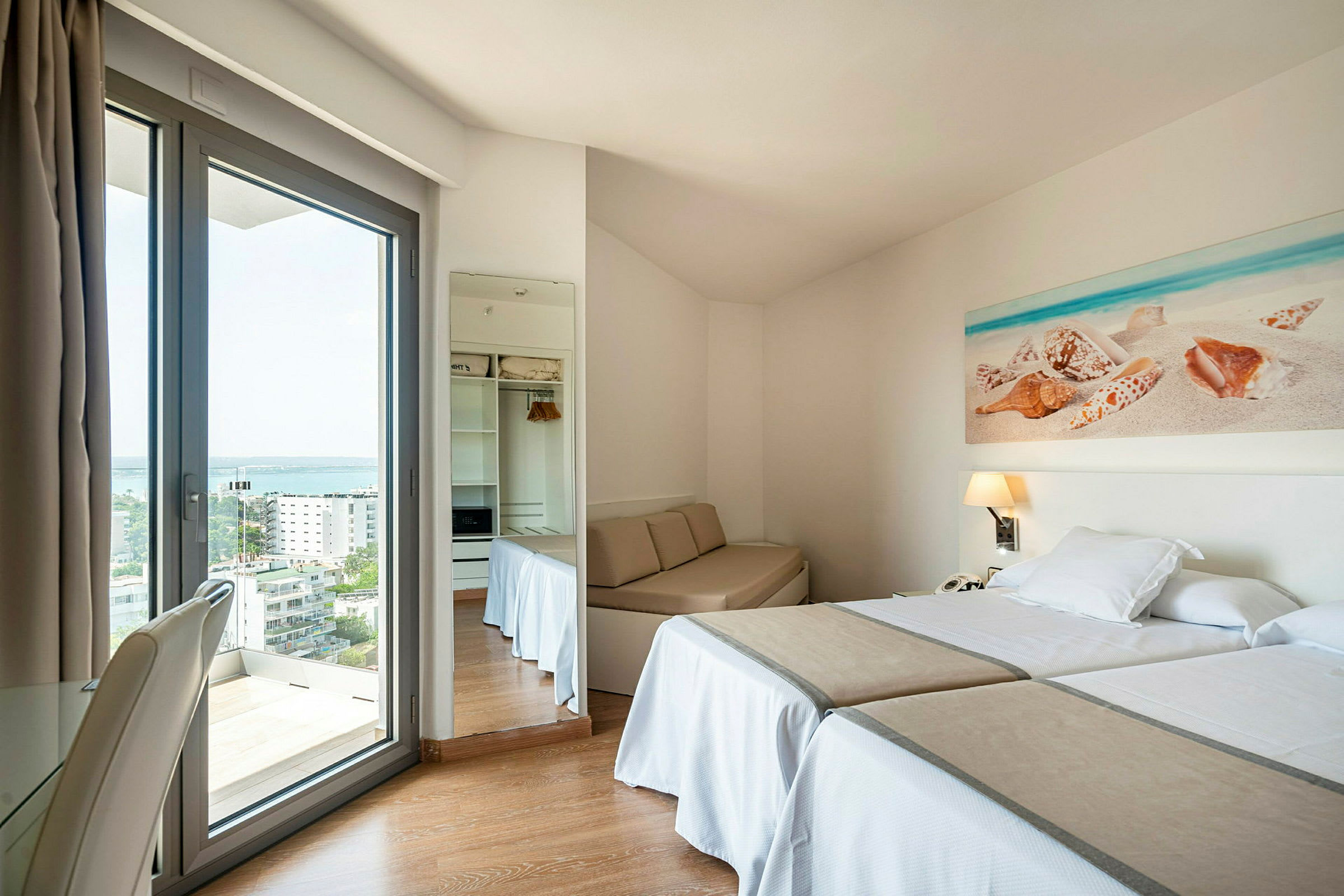 Hotellihuone, jossa kaksi yhden hengen vuodepaikkaa, pieni pöytäalue ja lasiovi näkymällä ulos. (Tekoälyn luoma teksti)