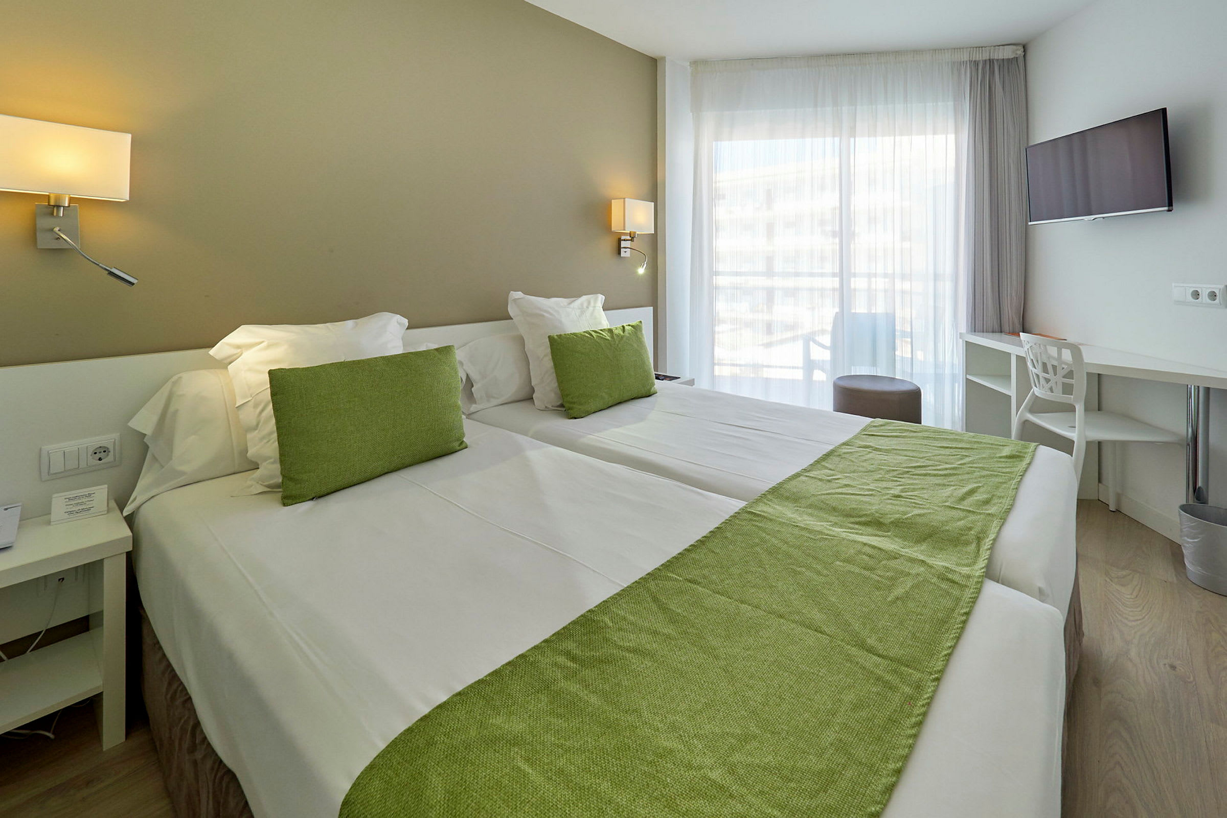 Hotellihuone kahdella vuoteella, vihreitä koristeita ja suuret ikkunat. (Tekoälyn luoma teksti)