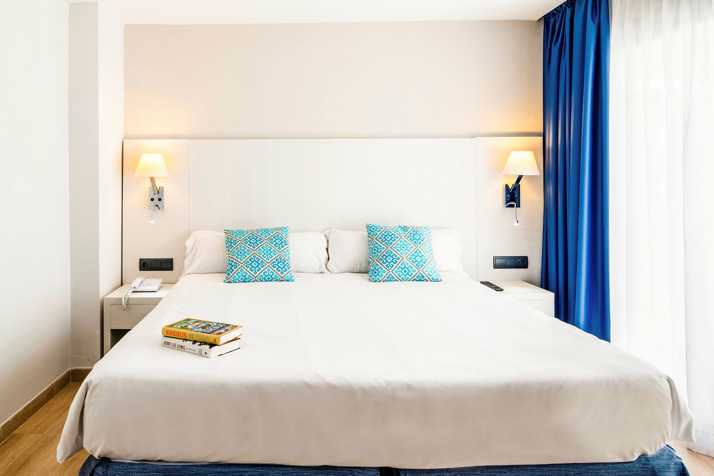 Moderni hotellihuone, jossa suuri parivuode, kaksi sinistä kuviollista koristetyynyä ja seinään kiinnitetyt lukulamput. Sängyn vieressä on yöpöytä ja ikkuna, jossa on valkoiset ja siniset verhot. (Tekoälyn luoma teksti)