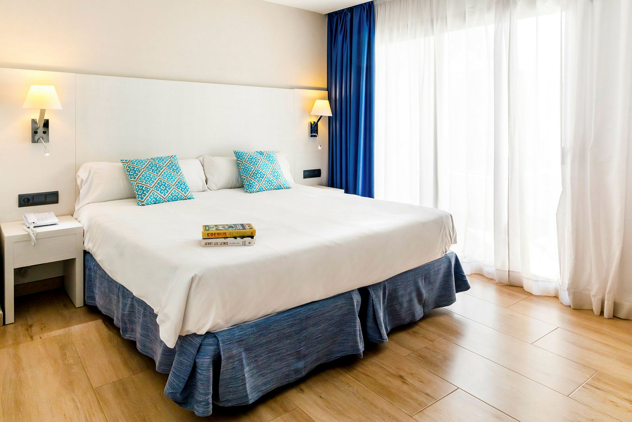 Hotellihuone, jossa on leveä sänky, valkoinen sängynpääty, siniset tyynyt ja verhot sekä vaalea puulattia. (Tekoälyn luoma teksti)