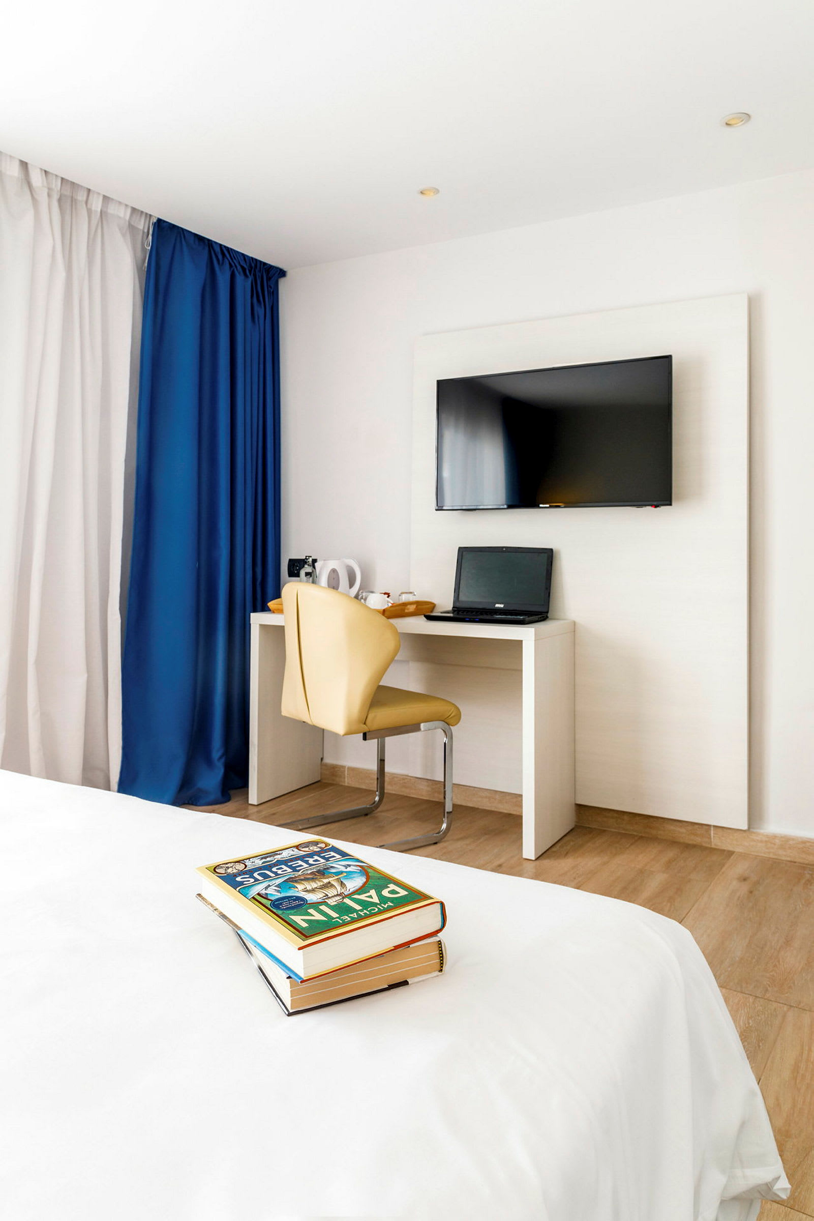 Hotellihuone, jossa seinällä on taulutelevisio, pöydällä kannettava tietokone ja beige tuoli. Etualalla näkyy sänky ja pino kirjoja. (Tekoälyn luoma teksti)