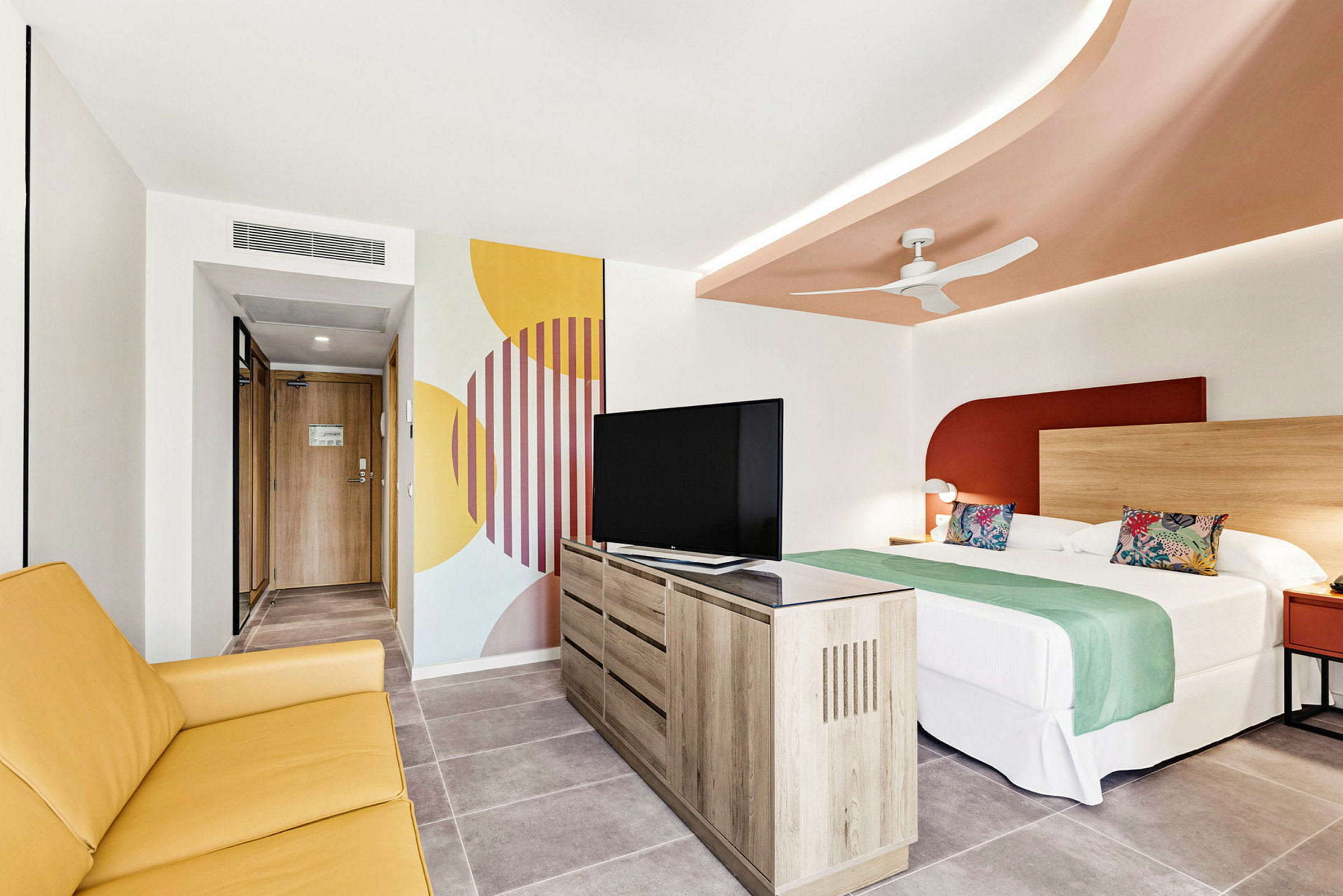 Hotellihuone, jossa on sänky, keltainen sohva, televisio ja koristeellinen seinämaalaus. (Tekoälyn luoma teksti)