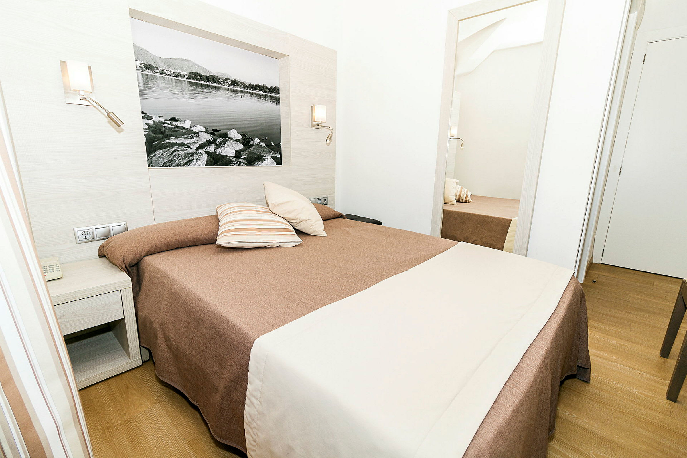 Hotellihuone, jossa on sänky ruskeilla ja valkoisilla vuodevaatteilla, yöpöytä vieressä ja peili, joka heijastaa huonetta. (Tekoälyn luoma teksti)