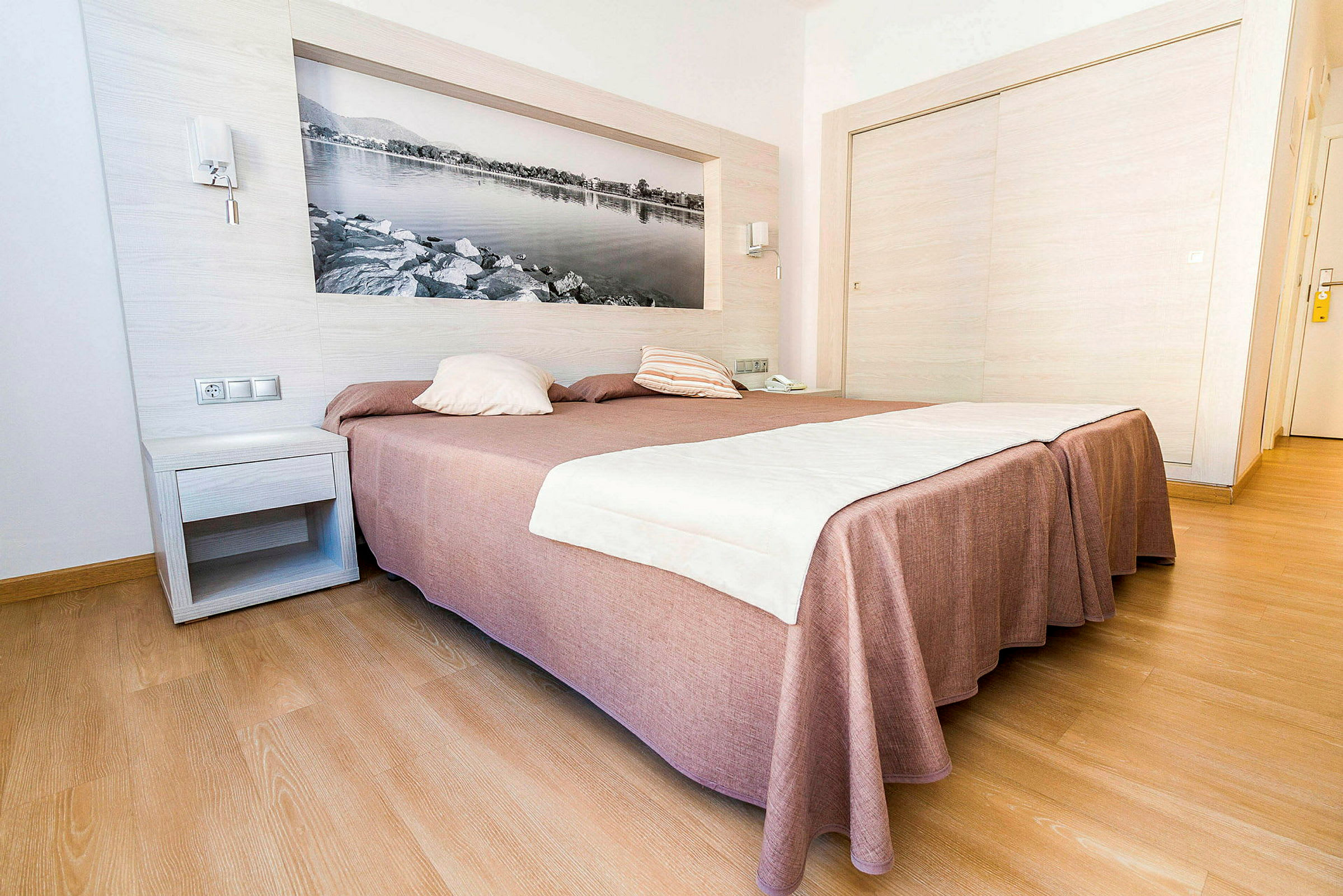 Hotellihuone, jossa on suuri sänky, ruskea päiväpeite ja valkoiset lakanat, puulattia ja seinäkoriste mustavalkoisen kuvan muodossa. (Tekoälyn luoma teksti)