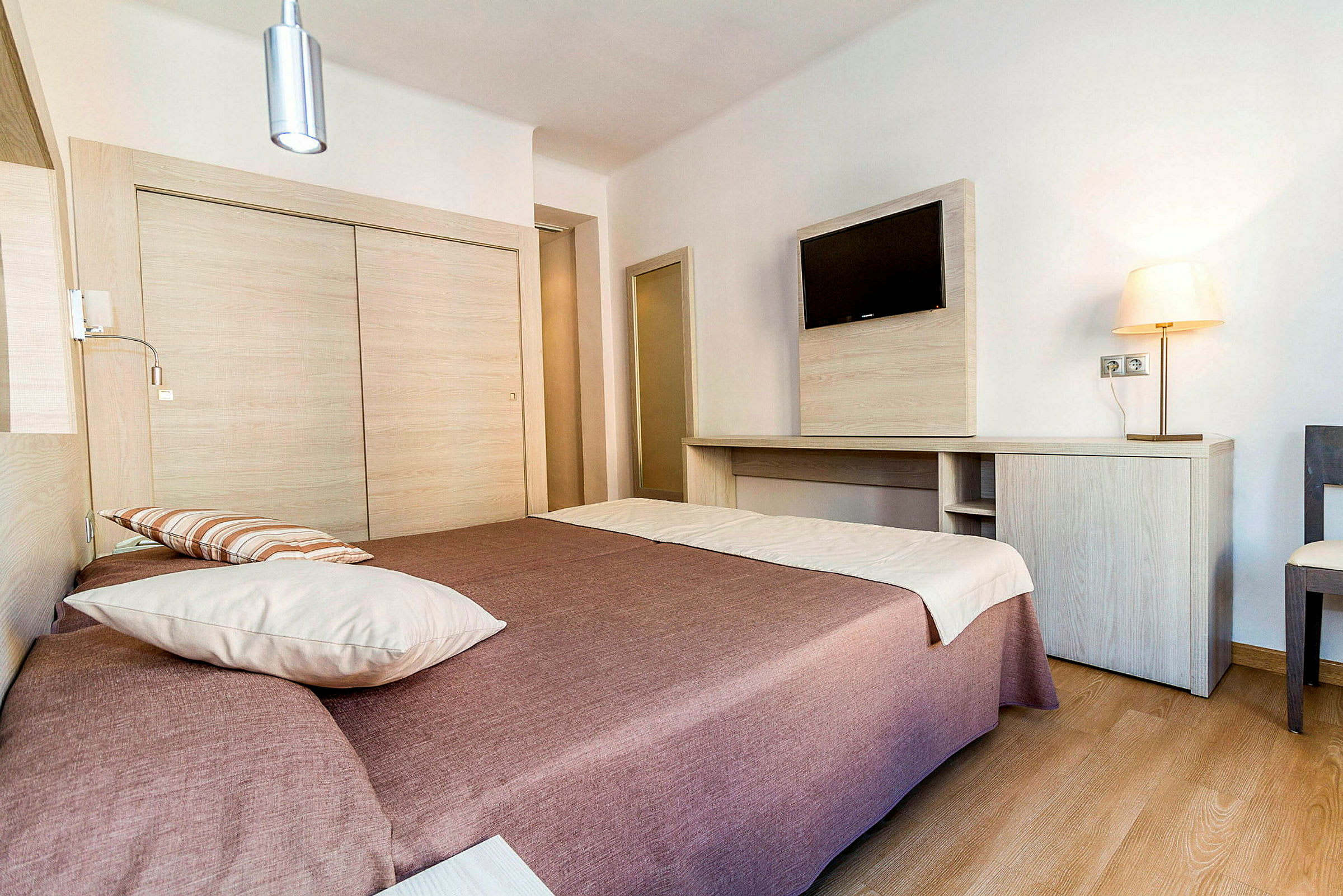 Hotellihuone, jossa on parisänky ruskealla päiväpeitteellä ja valkoisilla tyynyillä. Seinällä on taulu-tv puisen työpöydän yläpuolella. Huone on valoisa ja modernisti sisustettu. (Tekoälyn luoma teksti)
