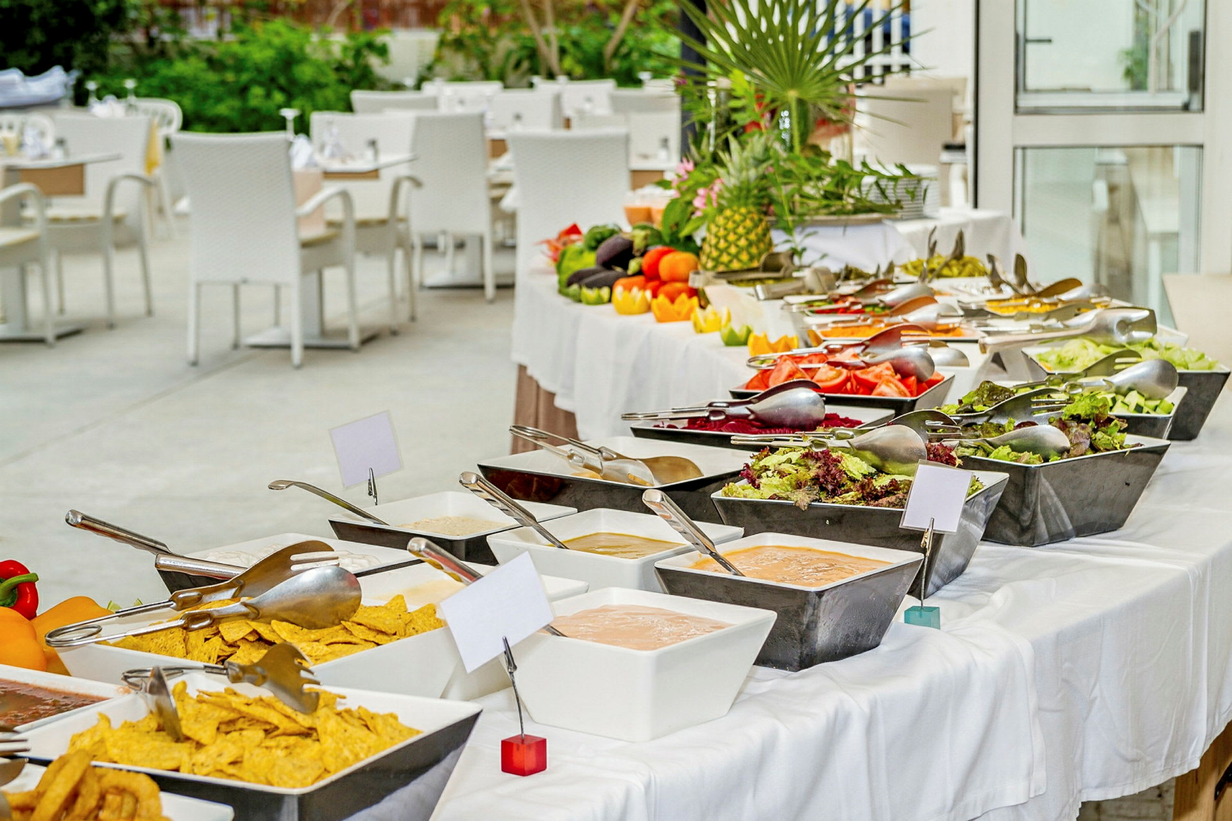 Buffet, jossa on salaattia, vihanneksia ja kastikkeita valkoisilla pöytäliinoilla olevalla pöydällä. Taustalla istumapaikkoja valkoisilla tuoleilla. (Tekoälyn luoma teksti)
