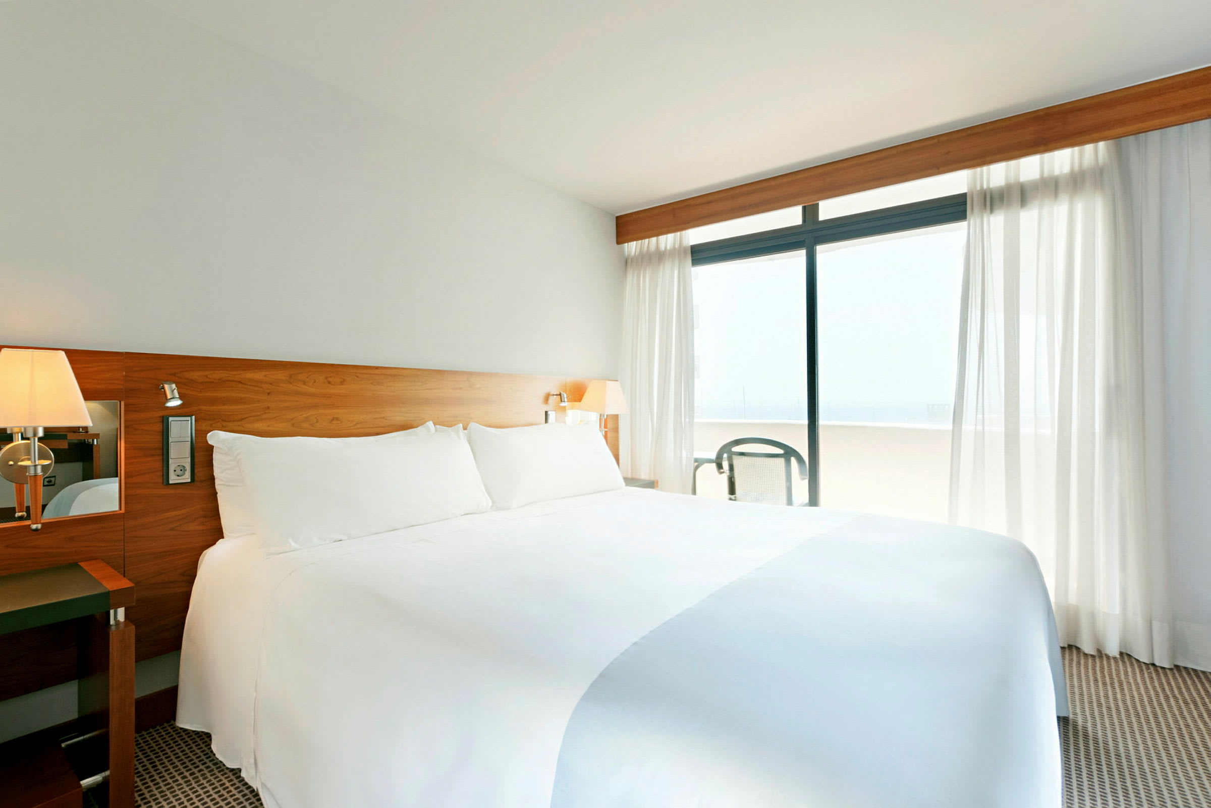Valoisa hotellihuone, jossa on suuri pedattu sänky ja valkoiset vuodevaatteet, puinen sängynpääty ja verhoilla varustetut ikkunat, jotka avautuvat parvekkeelle. (Tekoälyn luoma teksti)