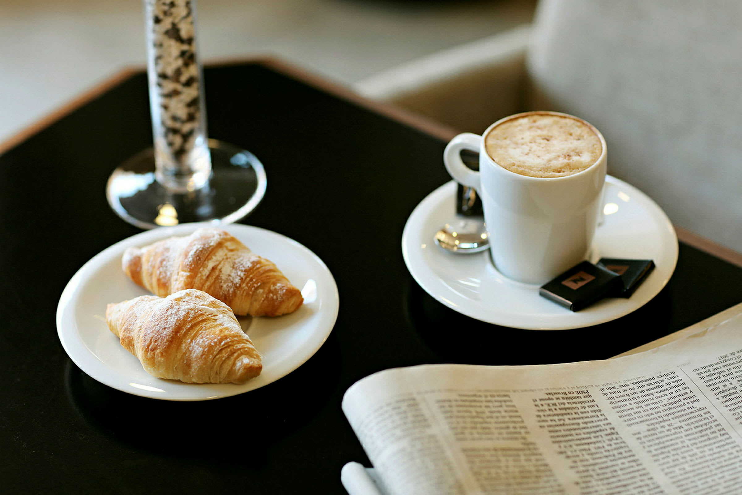 Kuvassa on kaksi croissantia lautasella, kupillinen maitokahvia vaahdolla ja lasi taustalla. Sanomalehti makaa pöydällä vieressä. (Tekoälyn luoma teksti)