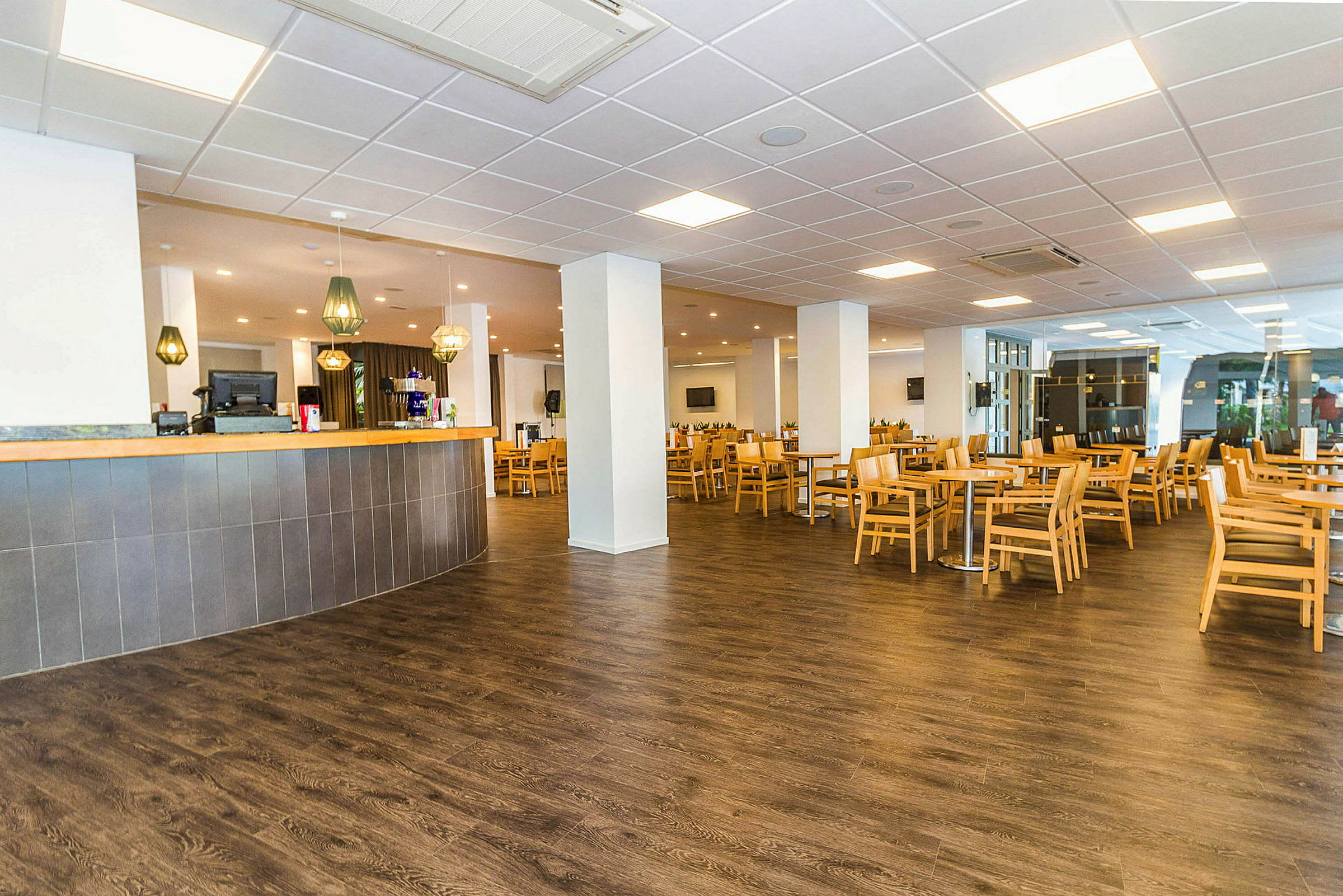 Tilava kahvila- tai ravintola-alue, jossa on vaaleita puupöytiä ja -tuoleja tummalla lattialla. Etualalla on baari, jonka yläpuolella roikkuu valaisimia. (Tekoälyn luoma teksti)