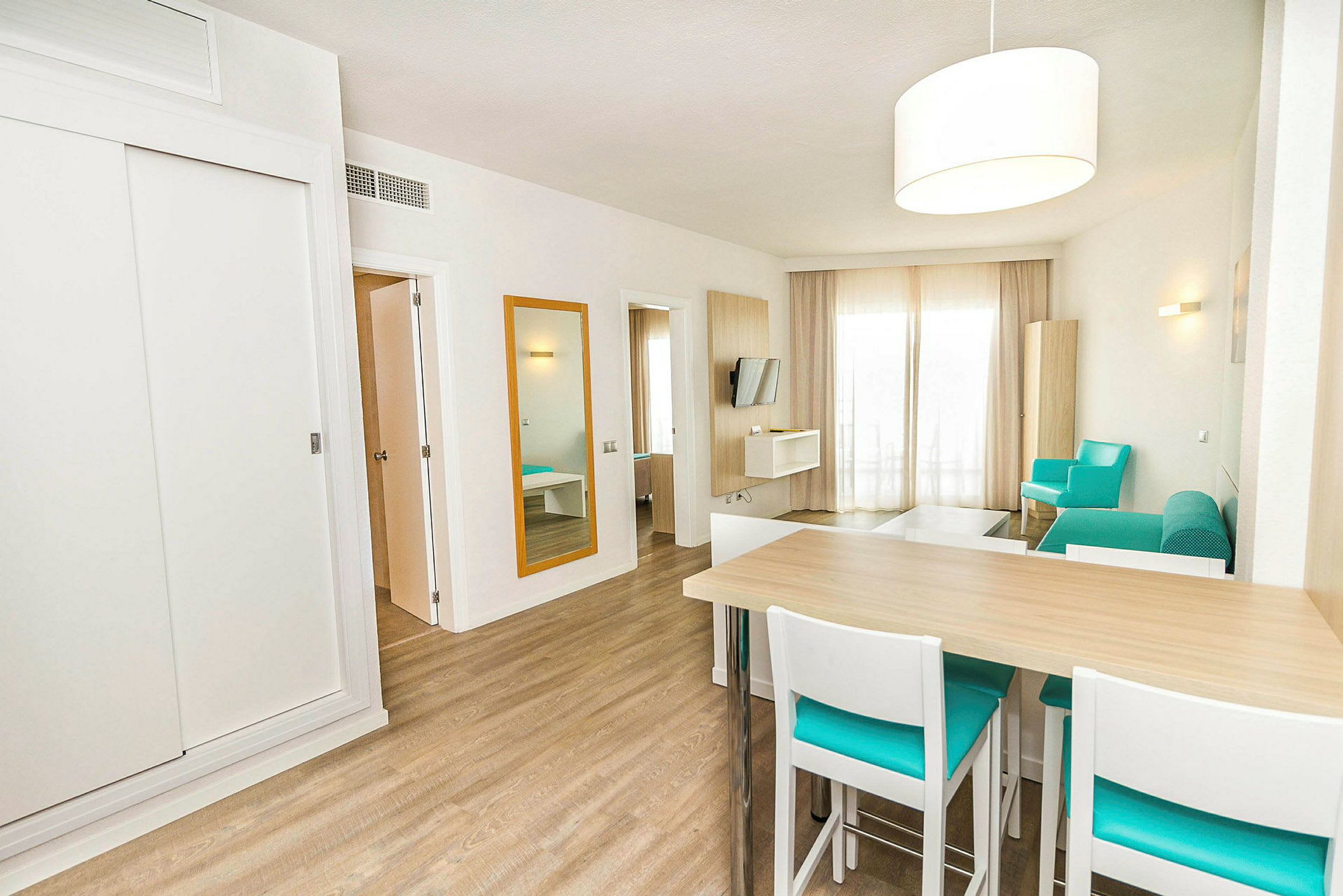 Vaaleasti ja modernisti sisustettu hotellihuone, jossa on puulattia, turkoosinvärisiä ja valkoisia huonekaluja sekä istuinsarja ja ruokapöytä. (Tekoälyn luoma teksti)