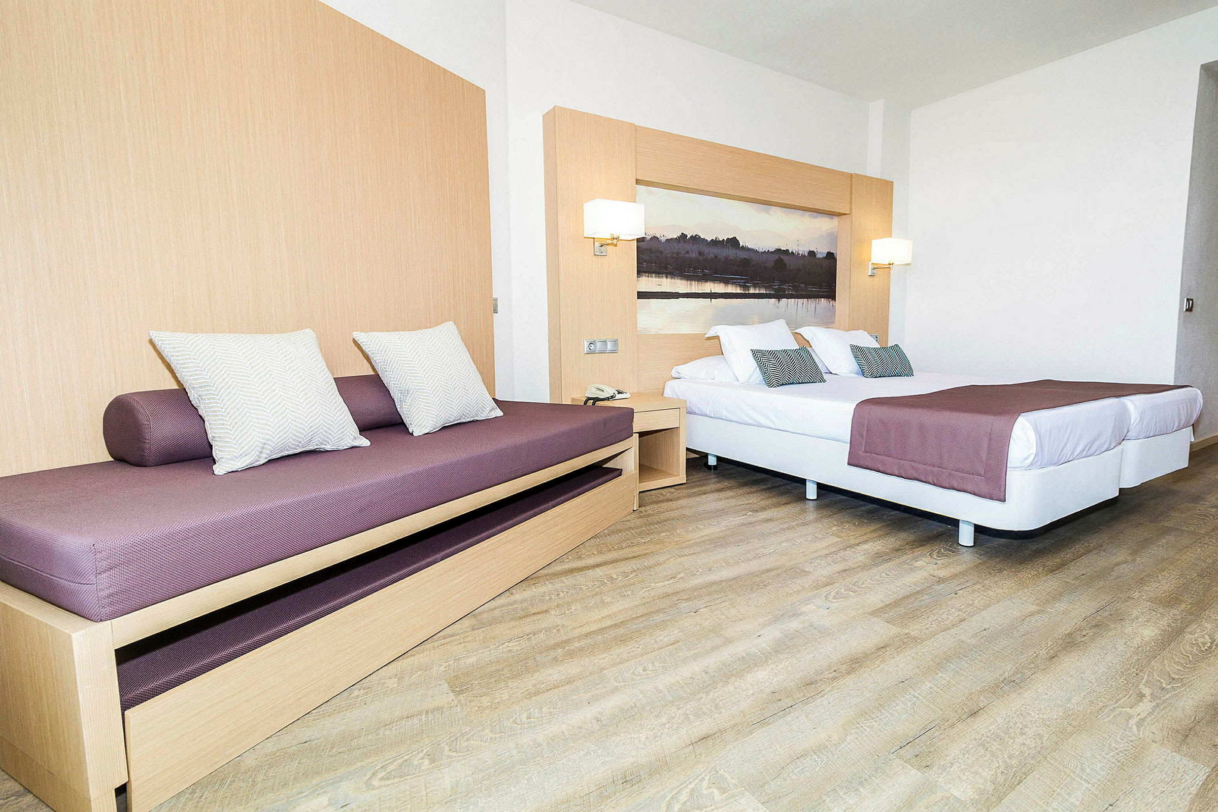 Hotellihuoneen sisustus, jossa vuodesohva ja parivuode, lilanväriset päiväpeitteet ja koristetyynyt. (Tekoälyn luoma teksti)