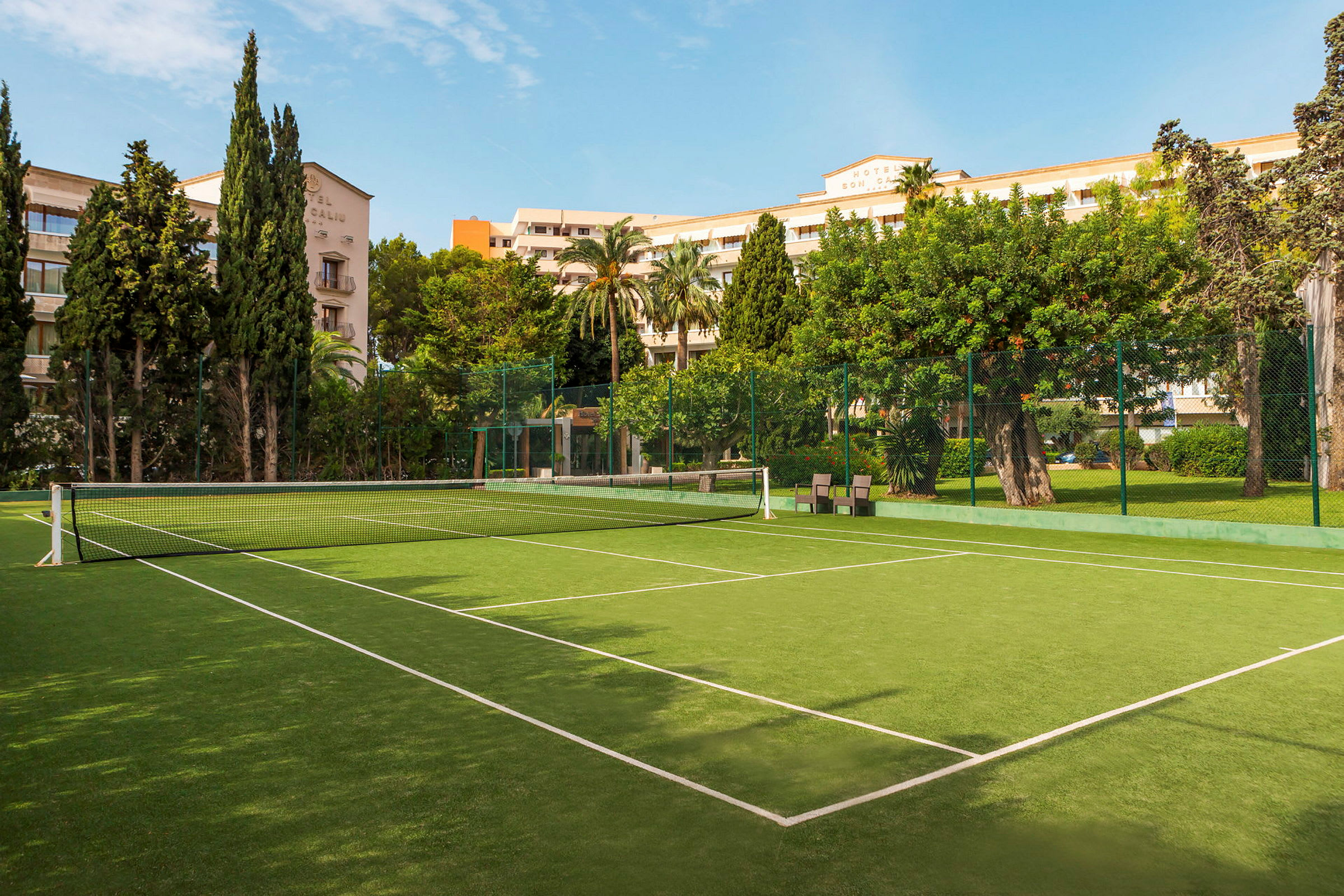 Tennis-kenttä vihreällä pinnalla ja valkoisilla linjoilla, ympäröitynä puilla ja rakennuksilla. (Tekoälyn luoma teksti)