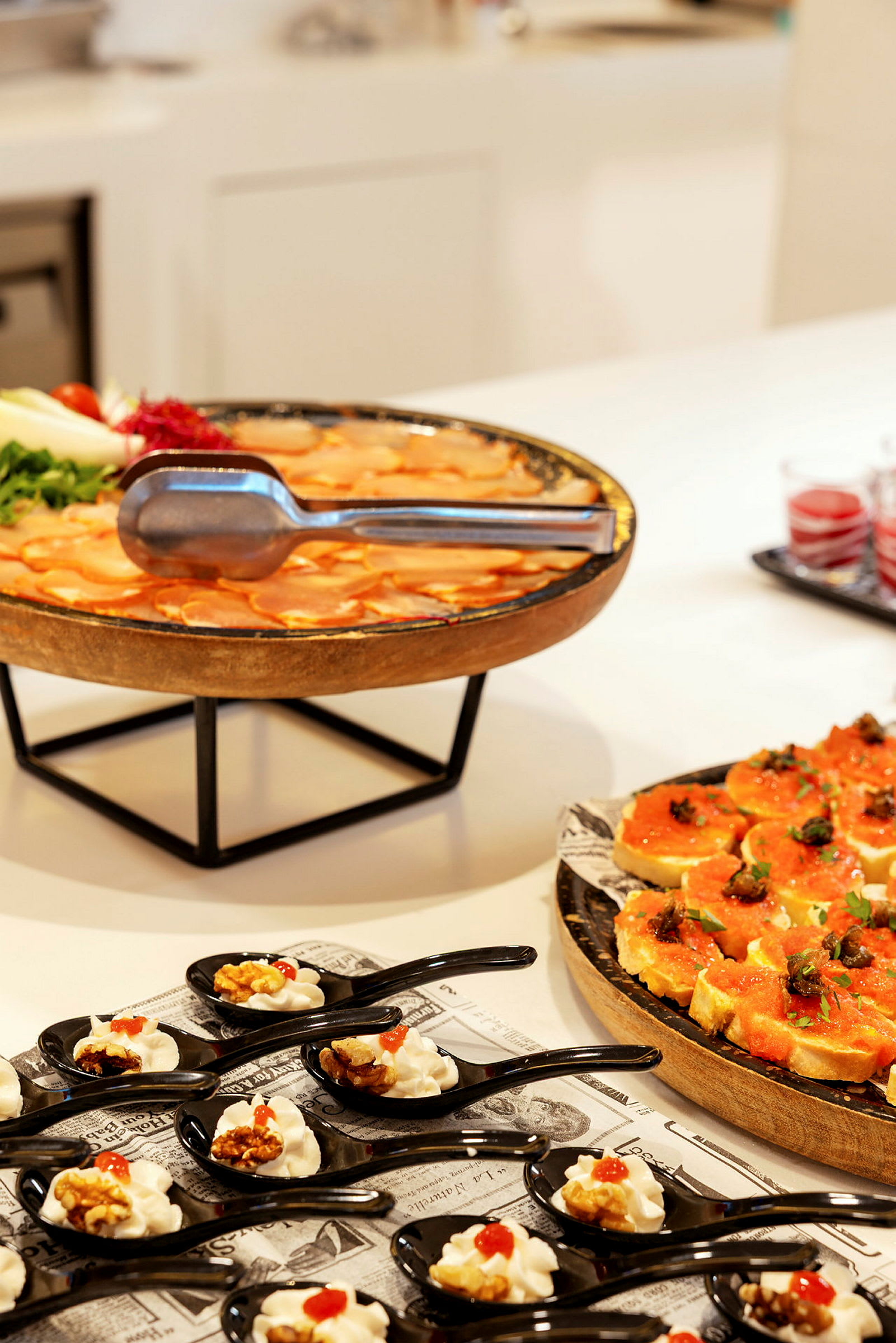 Buffet, jossa on erilaisia ruokia, mukaan lukien mustissa lusikoissa tarjoiltuja pieniä annoksia, oranssin värinen ruokalautasellinen ja koristeltuja leipäviipaleita. (Tekoälyn luoma teksti)