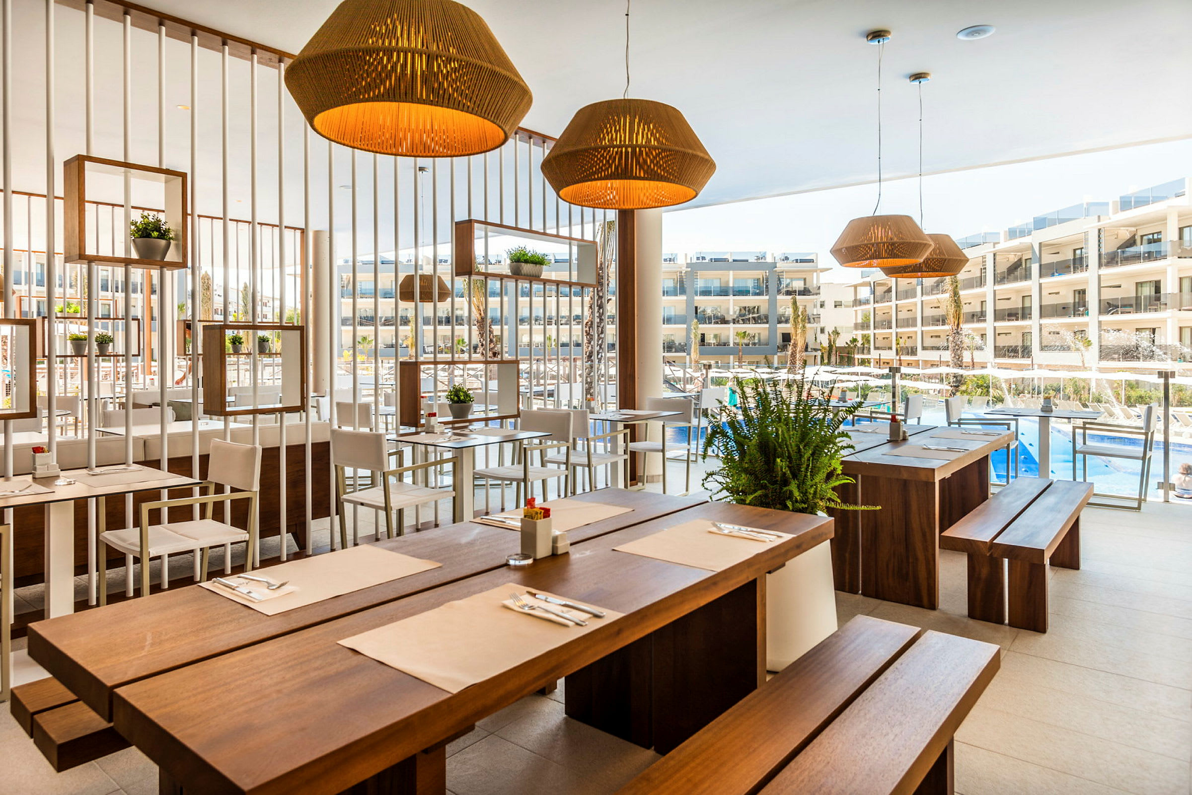Ravintola, jossa puiset pöydät ja vaaleat tuolit, koristeltu kasveilla ja lampuilla, näkymä uima-altaalle ja hotellirakennuksiin. (Tekoälyn luoma teksti)