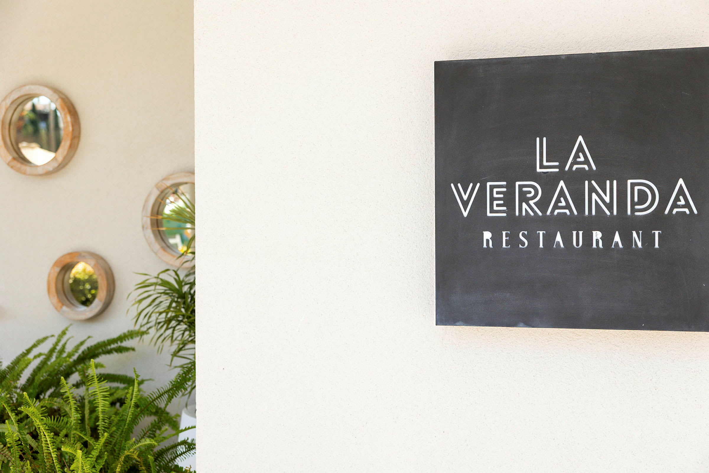 Kyltti, jossa lukee "La Veranda Restaurant", seinän vieressä, jossa on pyöreitä peilejä ja vihreitä kasveja. (Tekoälyn luoma teksti)