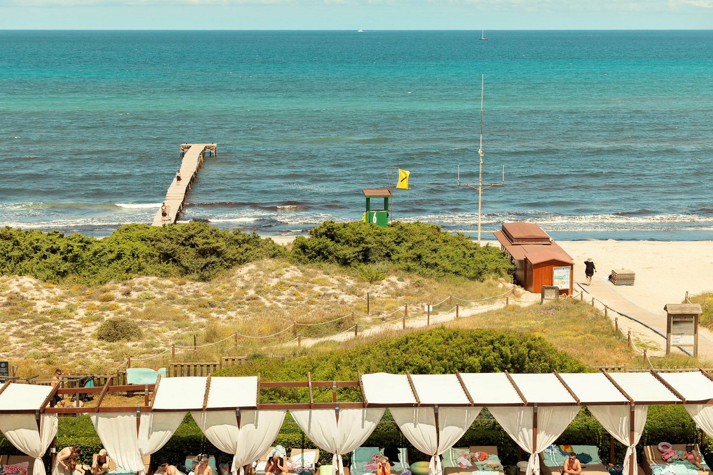 Ranta, josta laituri ulottuu mereen, hiekkadyynit kasvillisuudella etualalla ja ihmisiä rentoutumis- tai auringonottoalueella. (Tekoälyn luoma teksti)