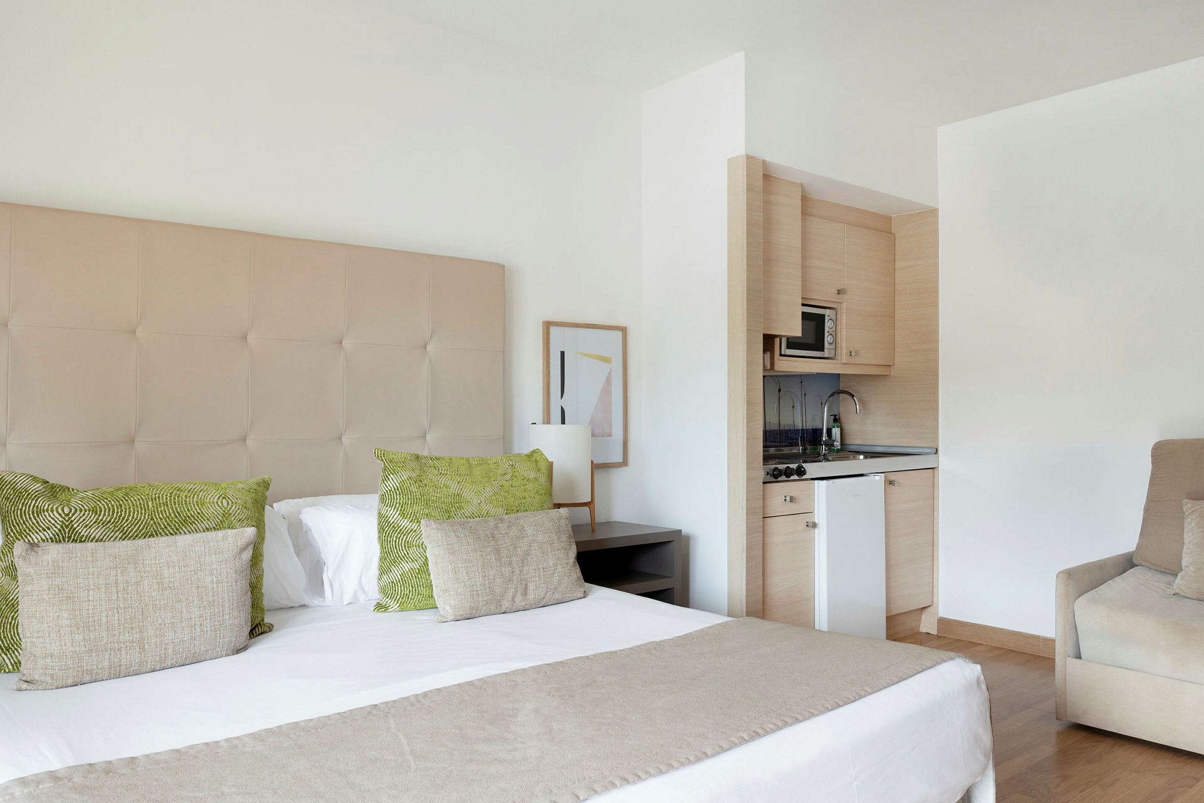 Hotellihuone, jossa on parivuode, vihreitä ja beigepäällisiä tyynyjä, pieni keittiötila mikroaaltouunin ja pesualtaan kanssa sekä beige nojatuoli. (Tekoälyn luoma teksti)