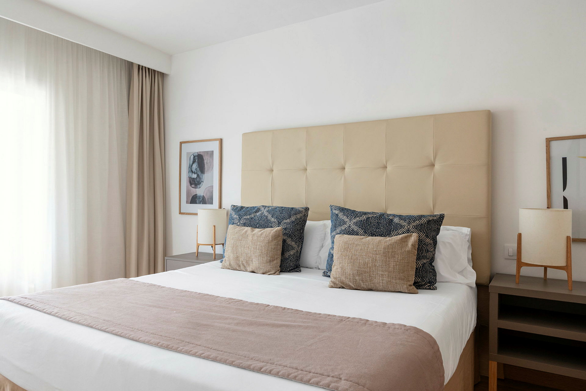 Hotellihuone, jossa on suuri sänky, peitetty valkoisilla lakanoilla, koristeellisilla tyynyillä ja vaalealla peitteellä. Sänky on sijoitettu kahden yöpöydän väliin, joissa on lamput. (Tekoälyn luoma teksti)