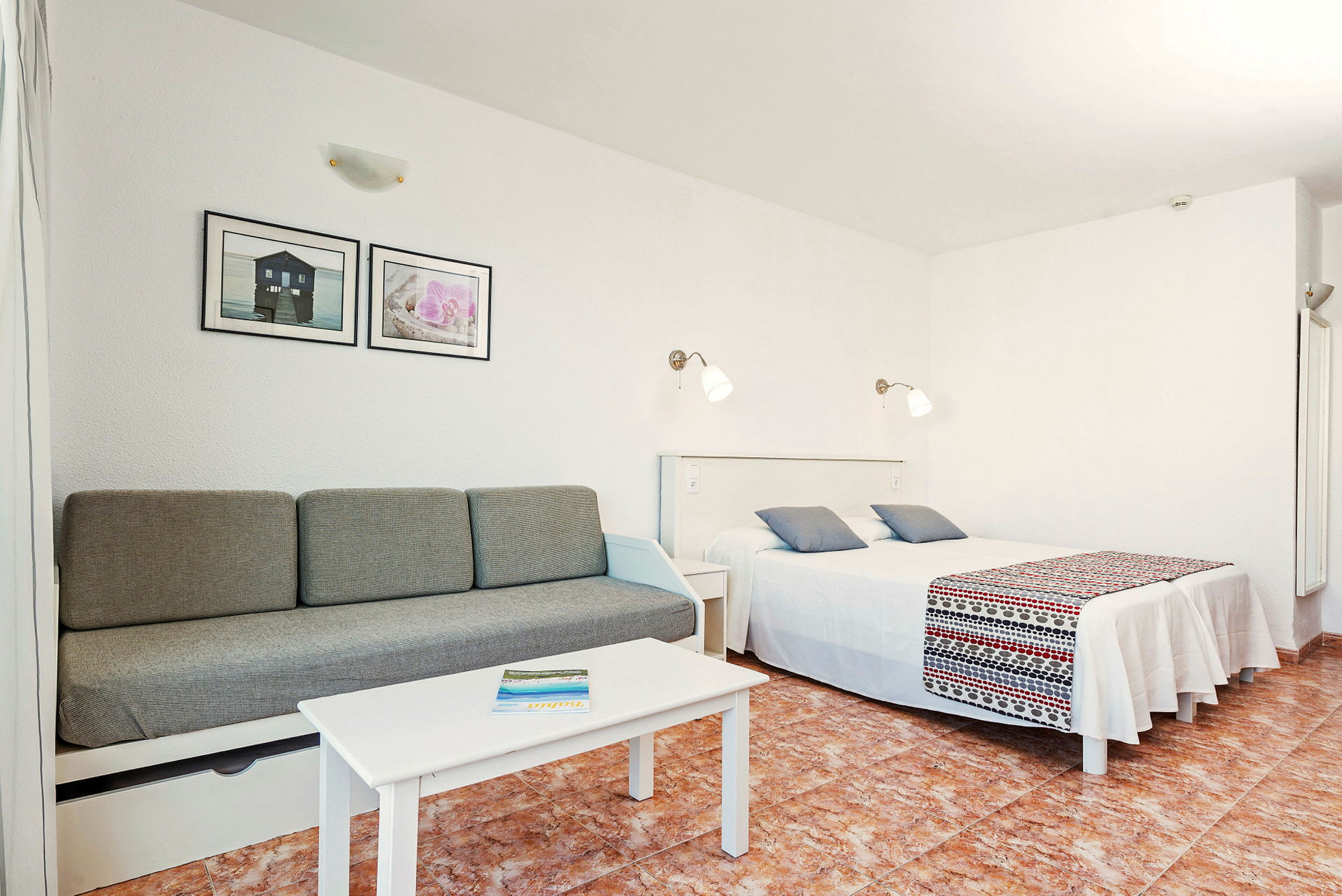 Hotellihuone, jossa vuodesohva, pöytä ja parivuode. Seinät ovat valkoiset, ja sohvan yläpuolella roikkuu tauluja. (Tekoälyn luoma teksti)