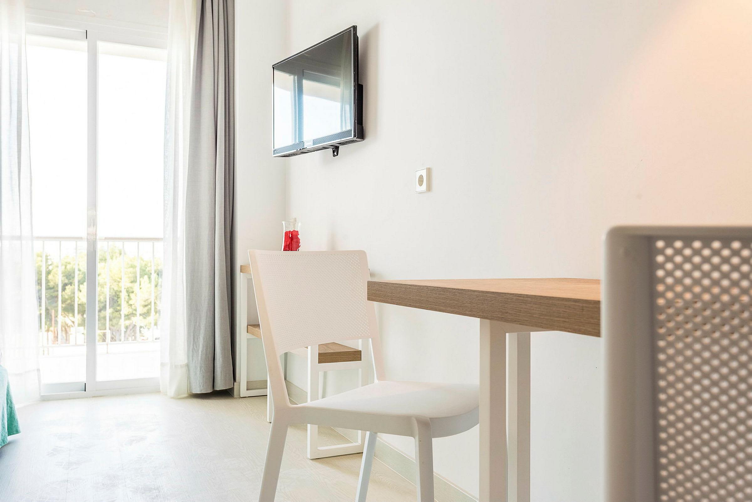 Hotellihuone, jossa valkoinen sisustus, pöytä ja tuolit sekä seinälle asennettu televisio. Taustalla lasiovet, jotka johtavat parvekkeelle ja näkymä viheralueelle. (Tekoälyn luoma teksti)