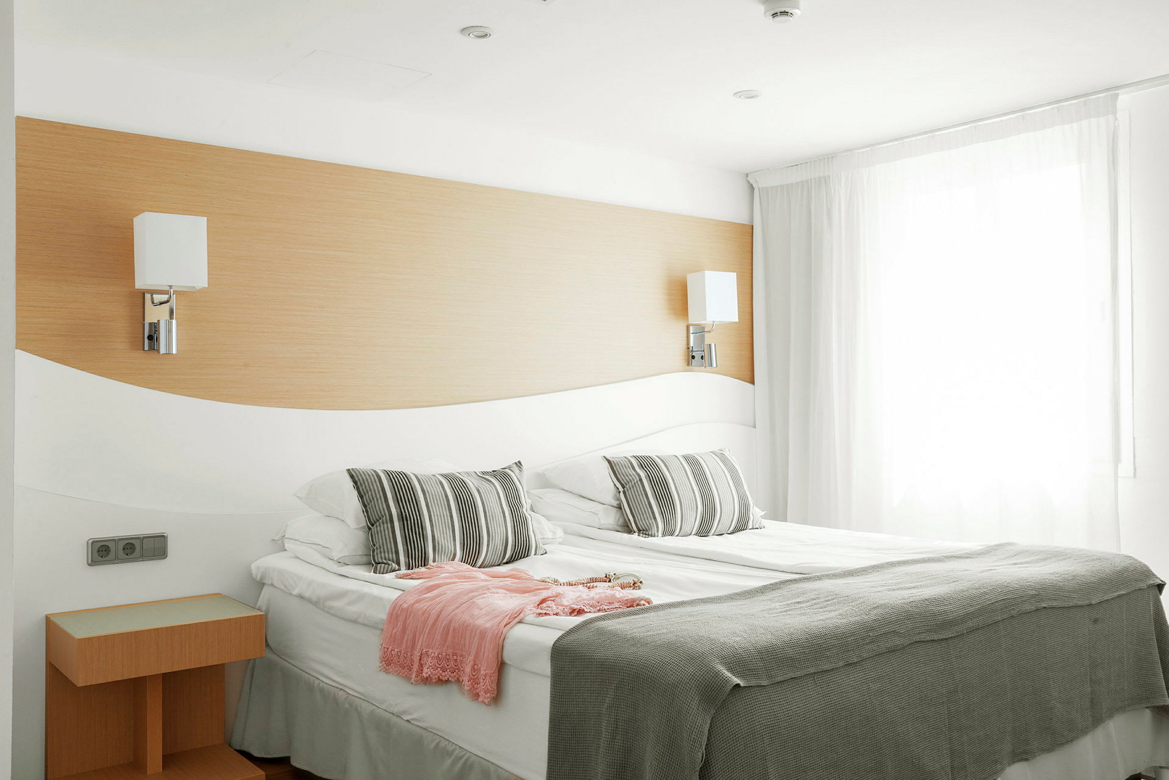 Hotellihuone, jossa on parivuode, kaksi lamppua seinällä ja harmaa sekä valkoinen peitto sekä tyynyt. (Tekoälyn luoma teksti)